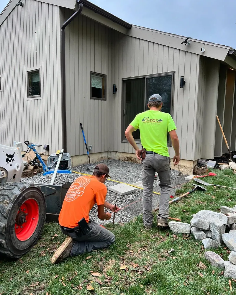 Two men are working on a patio in front of a house.