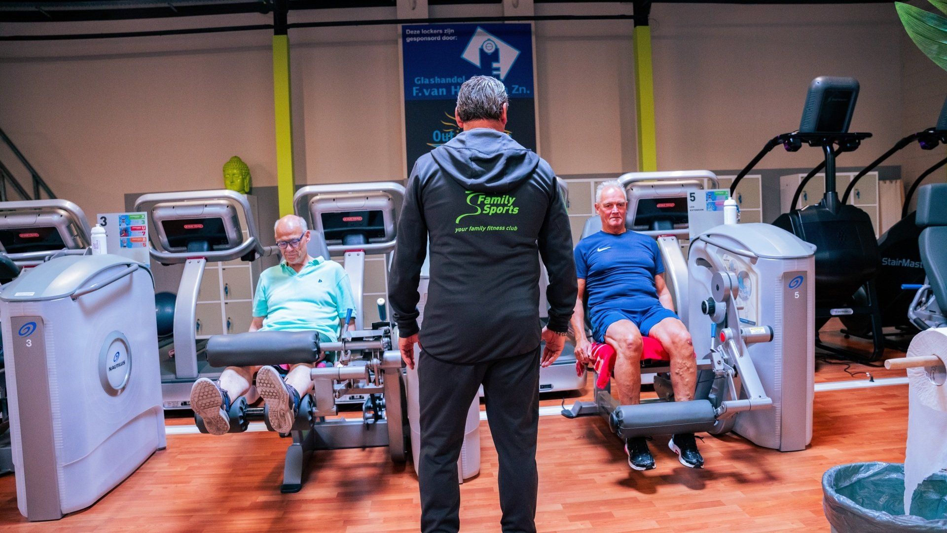 Een man staat voor een groep mensen die op fitnessapparaten in een sportschool zitten.