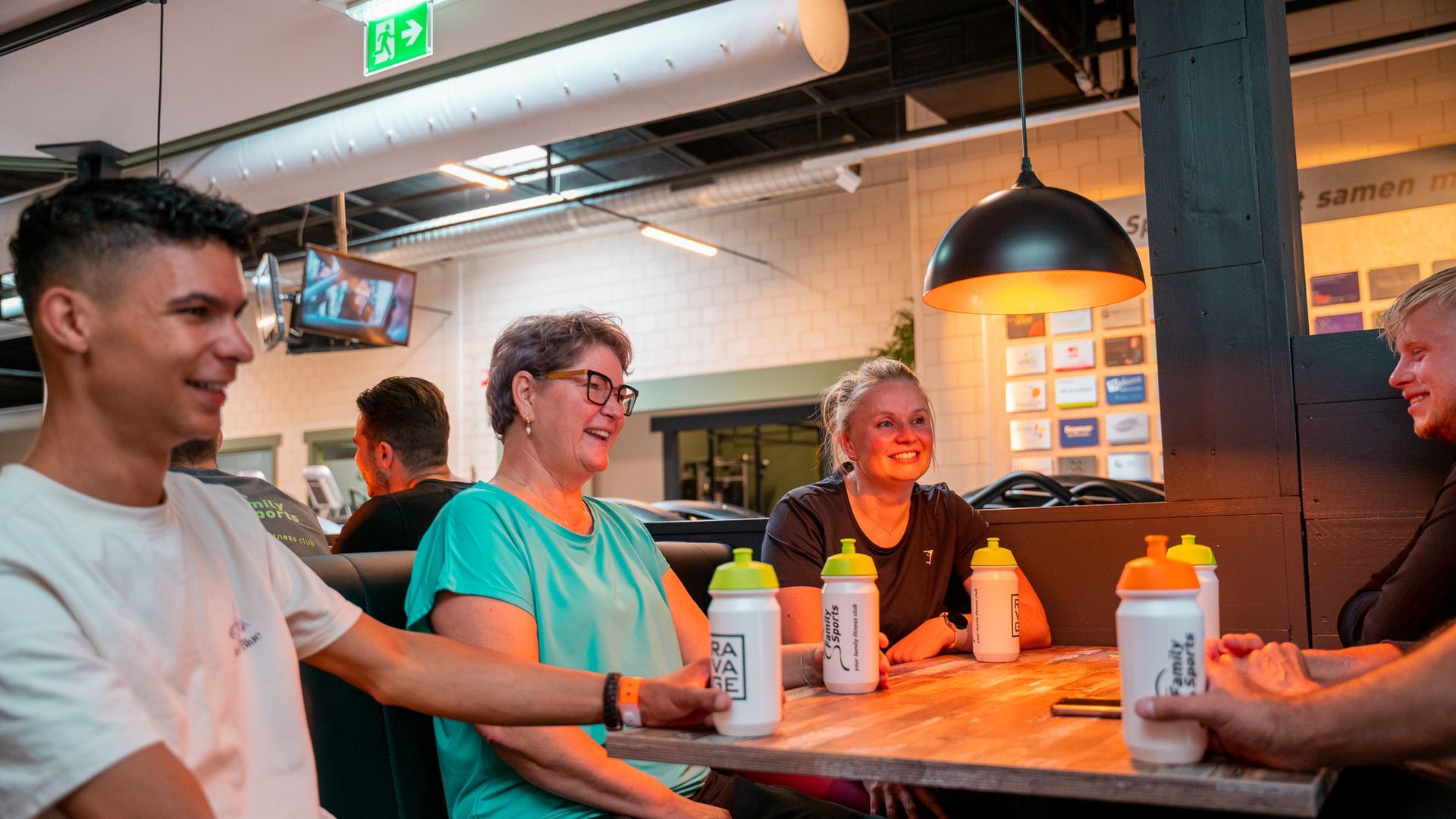 Vier mensen zitten aan een tafel in een gymzaal, lachend en pratend, met waterflesjes in hun handen.