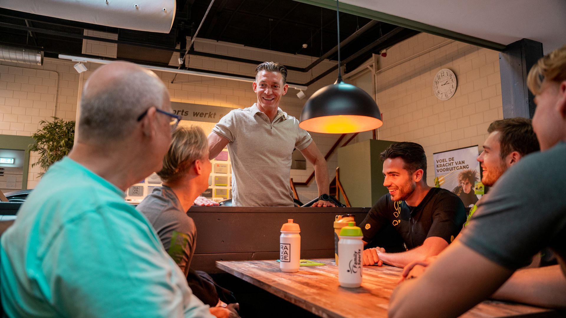 Mensen verzameld rond een tafel; een man staat er glimlachend bij. Interieur met waterflesjes.