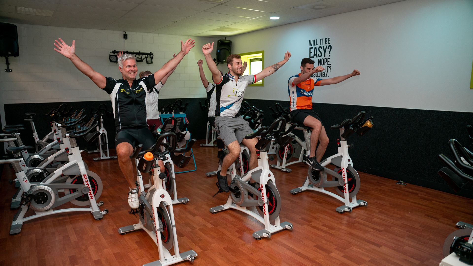 Drie mensen vieren hun vreugde op hometrainers in een sportschool, met hun armen omhoog.