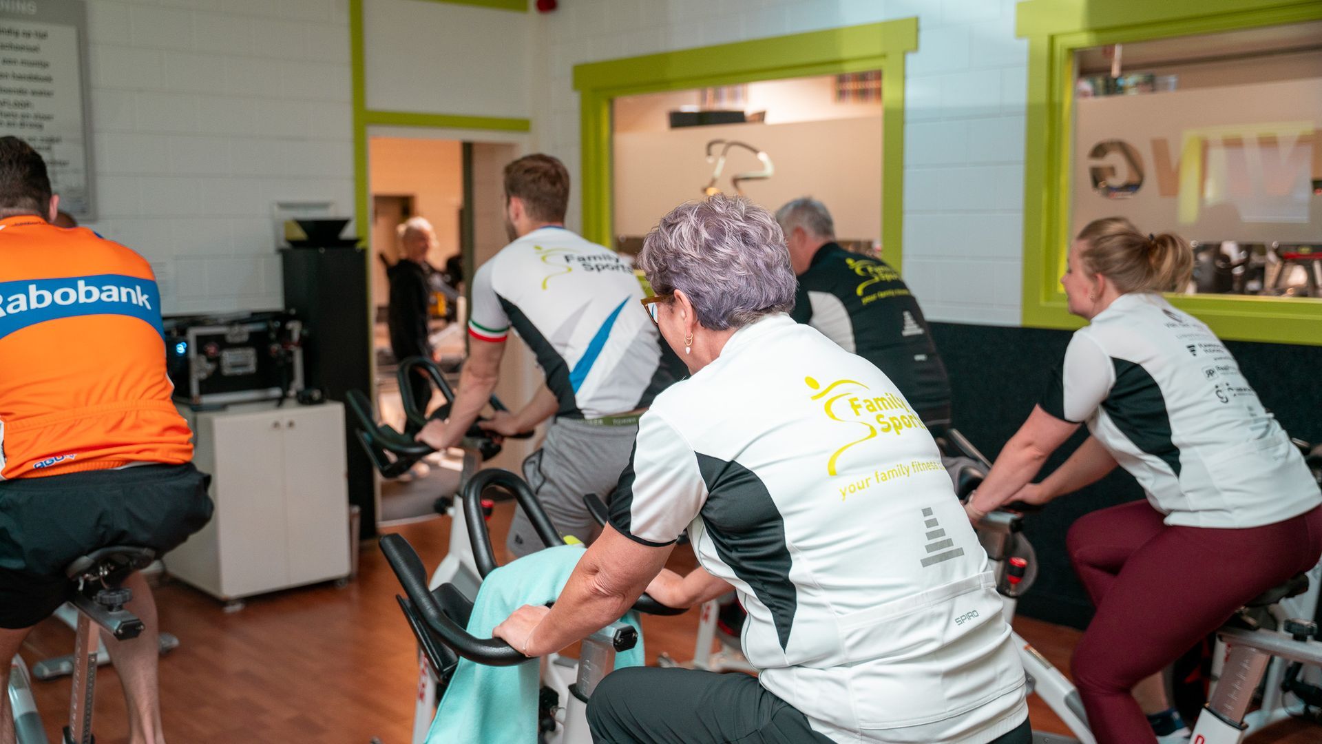 Mensen fietsen binnen in een sportschool; sommigen dragen wielershirts.