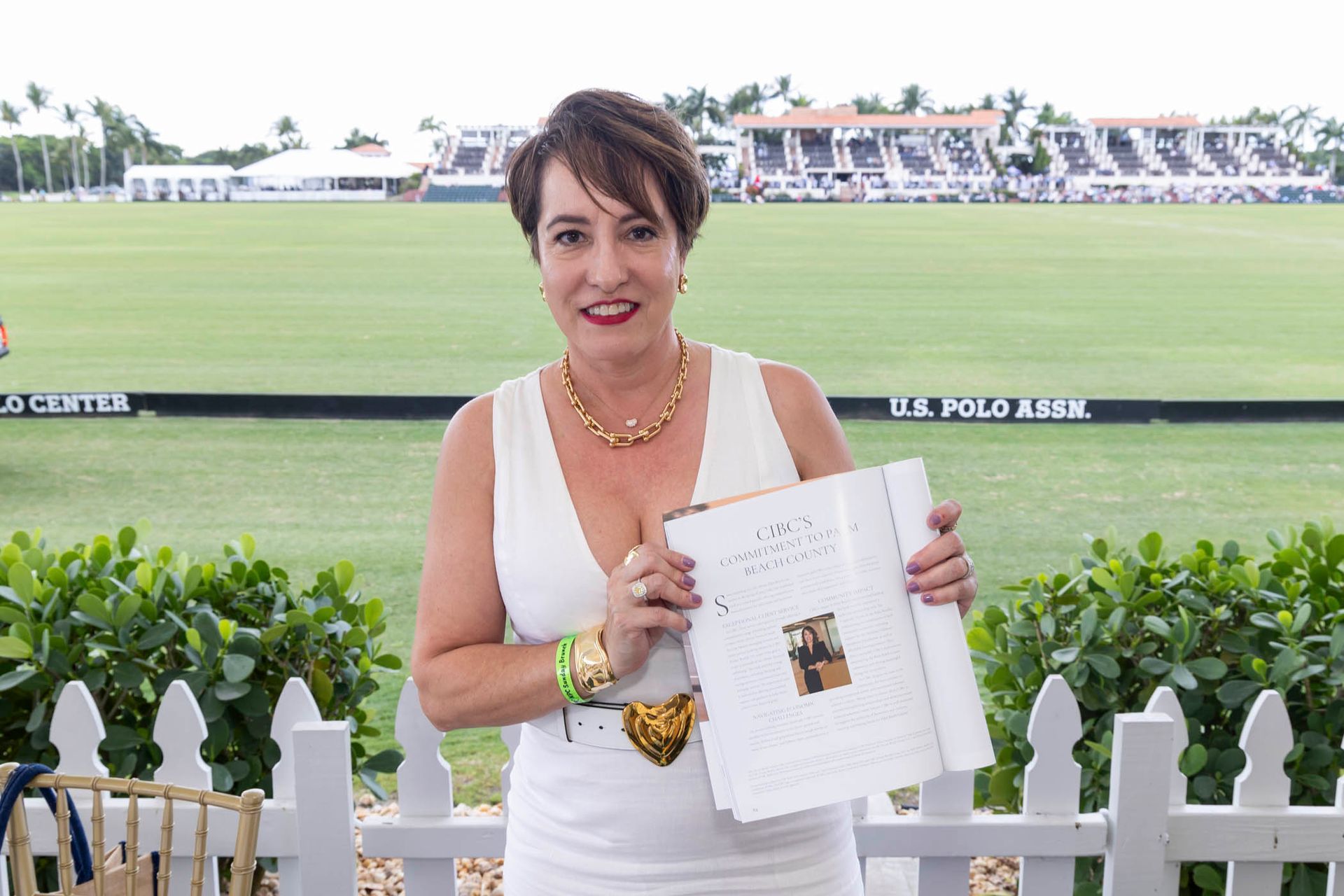 A woman in a white dress is holding a book in front of a polo field.