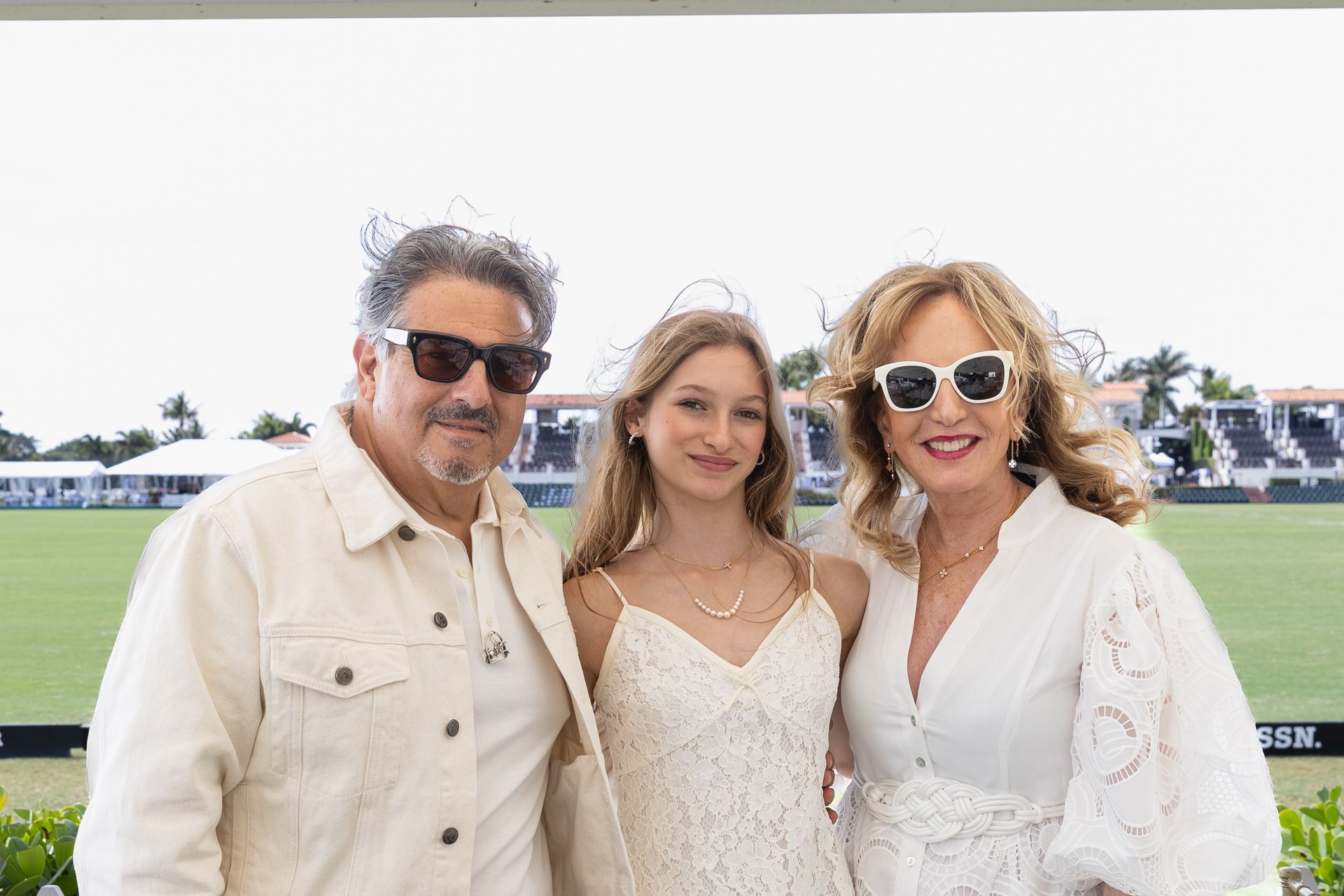 Family posing outdoors at a polo match. Man and woman in sunglasses, young woman in a dress, all in light colors.