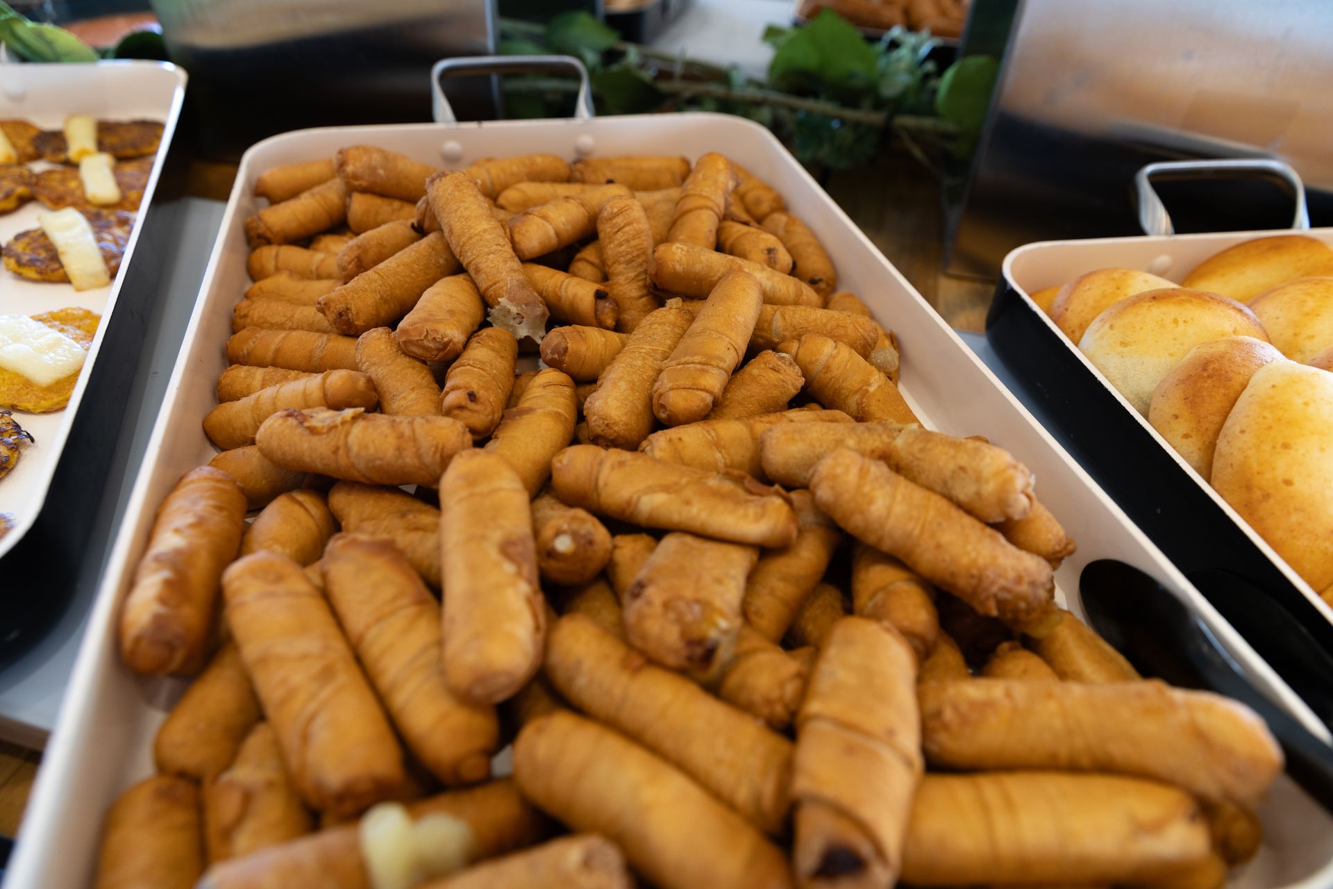 Tray of golden-brown tequeños, a fried cheese pastry, at a buffet with other food.