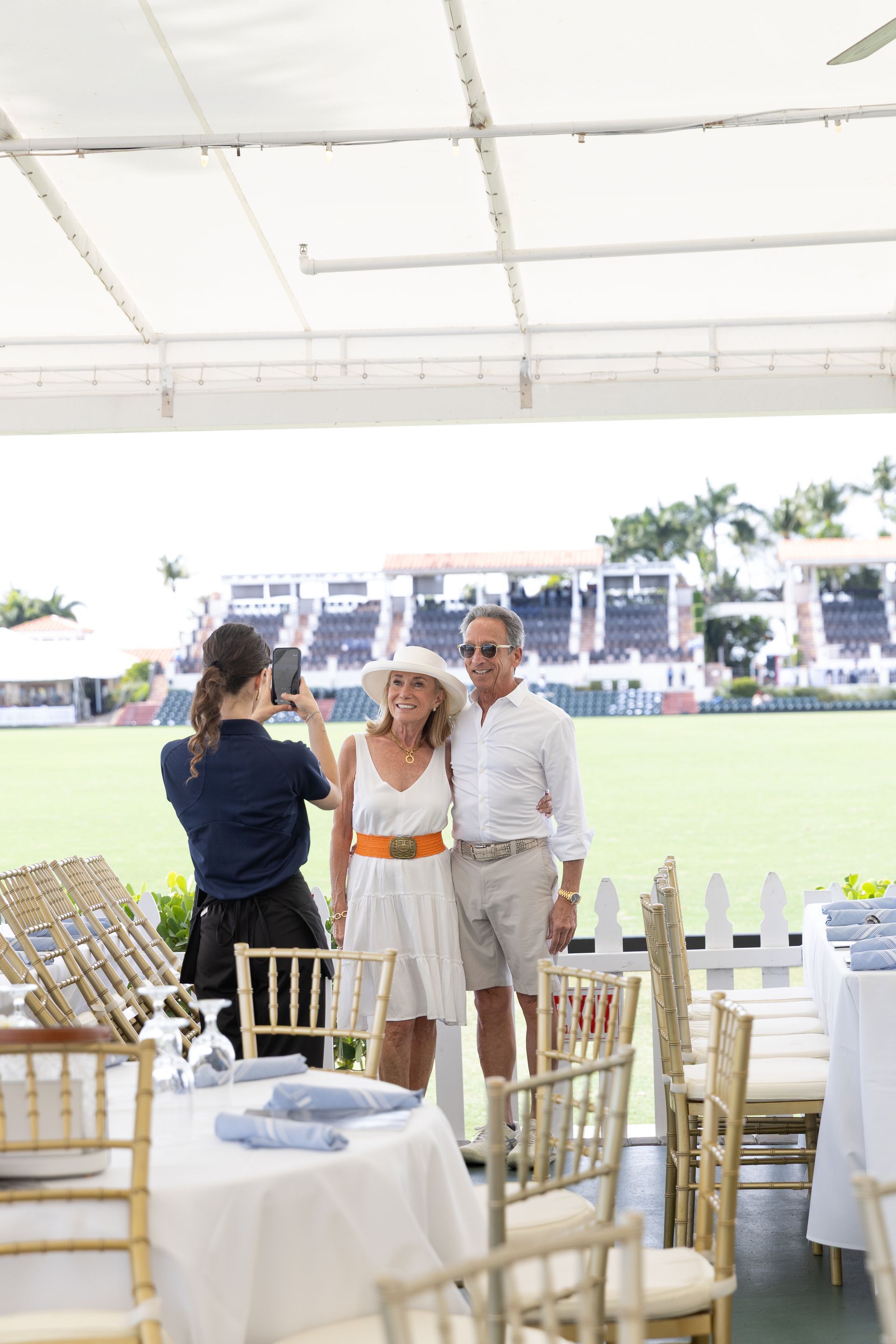 Woman taking photo of a couple posing. They stand in front of a polo field; he wears shorts and she a white dress.