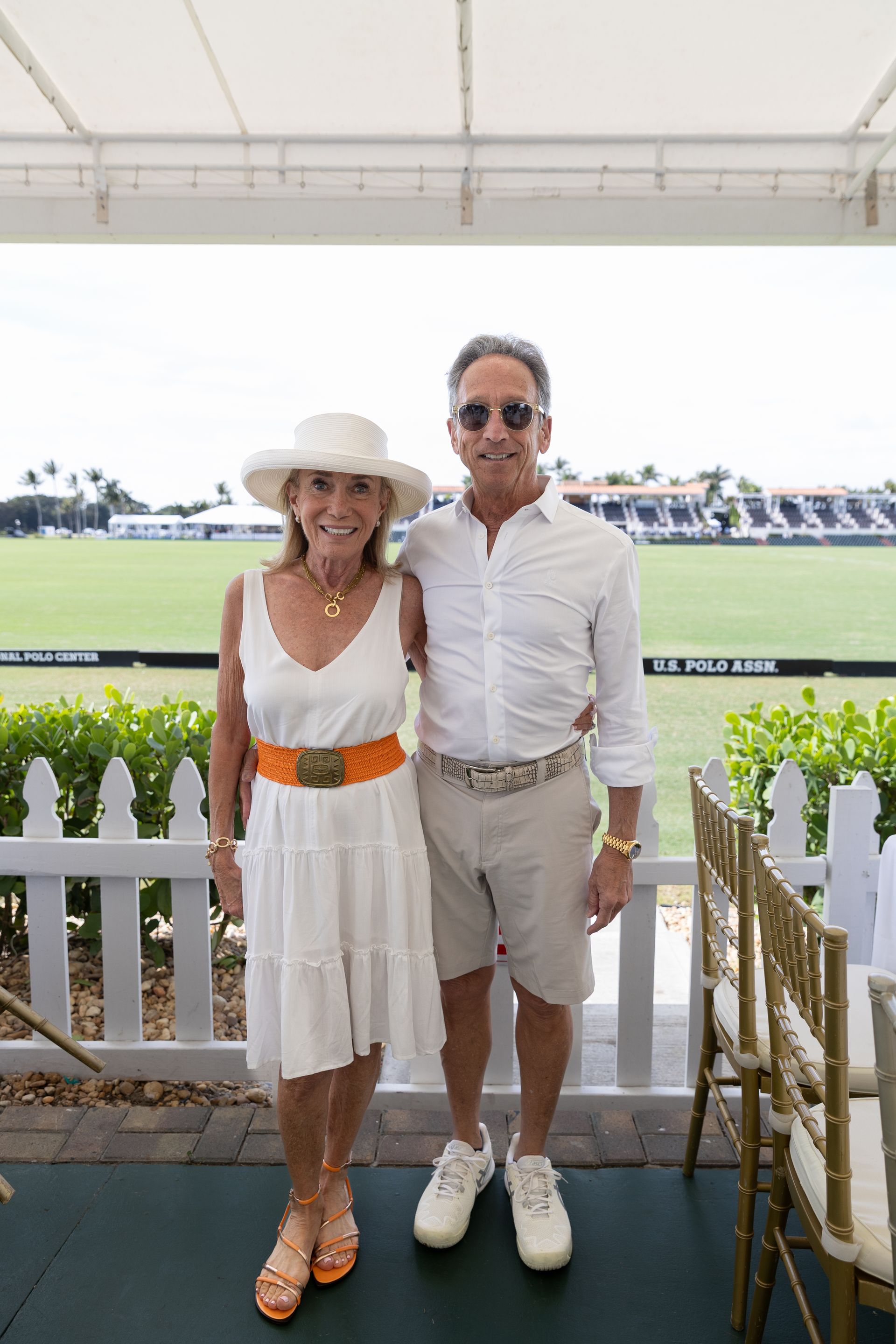 Couple in white attire at polo event, posing by a white fence.