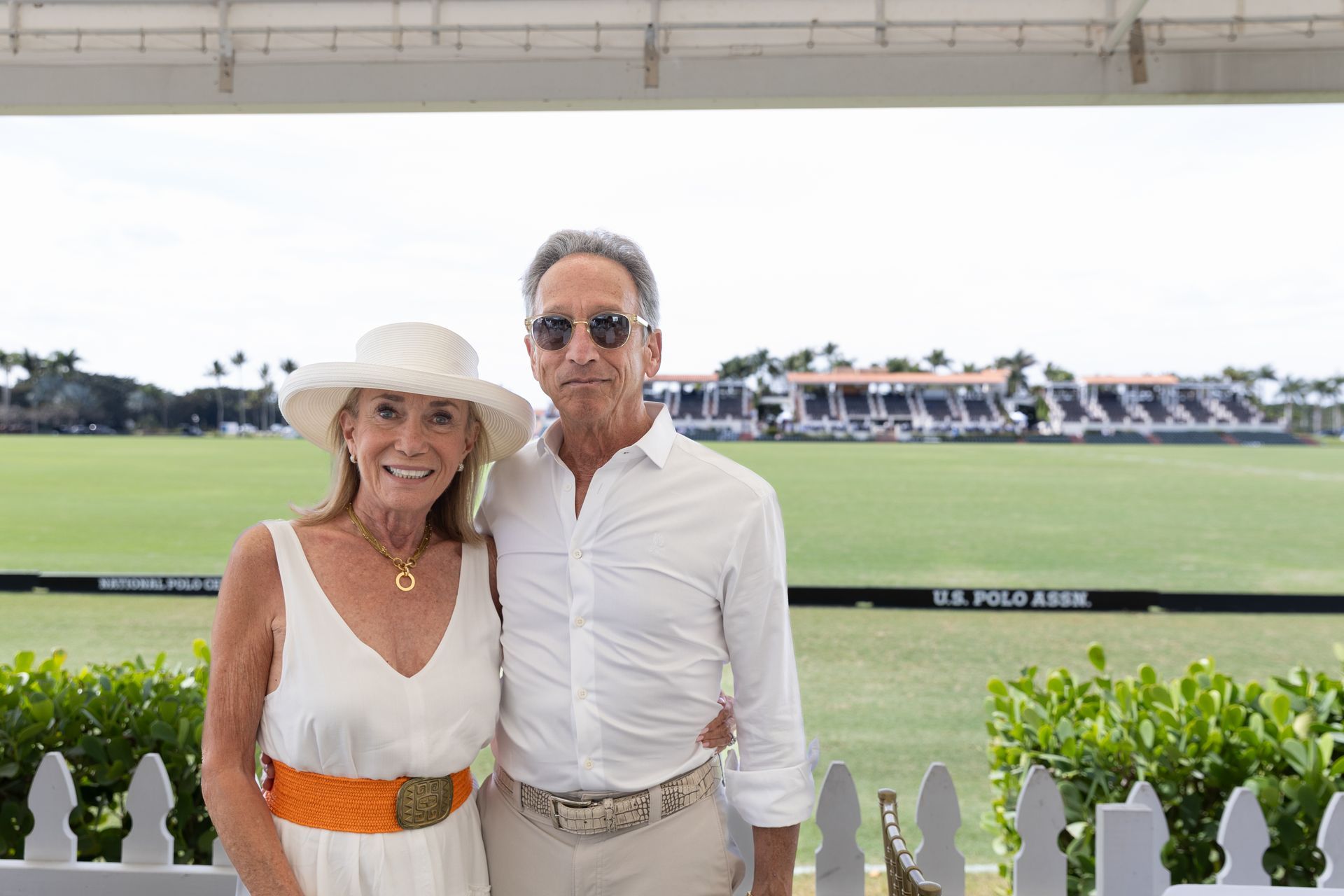 Woman in white dress and hat, man in white shirt, standing, polo field background.