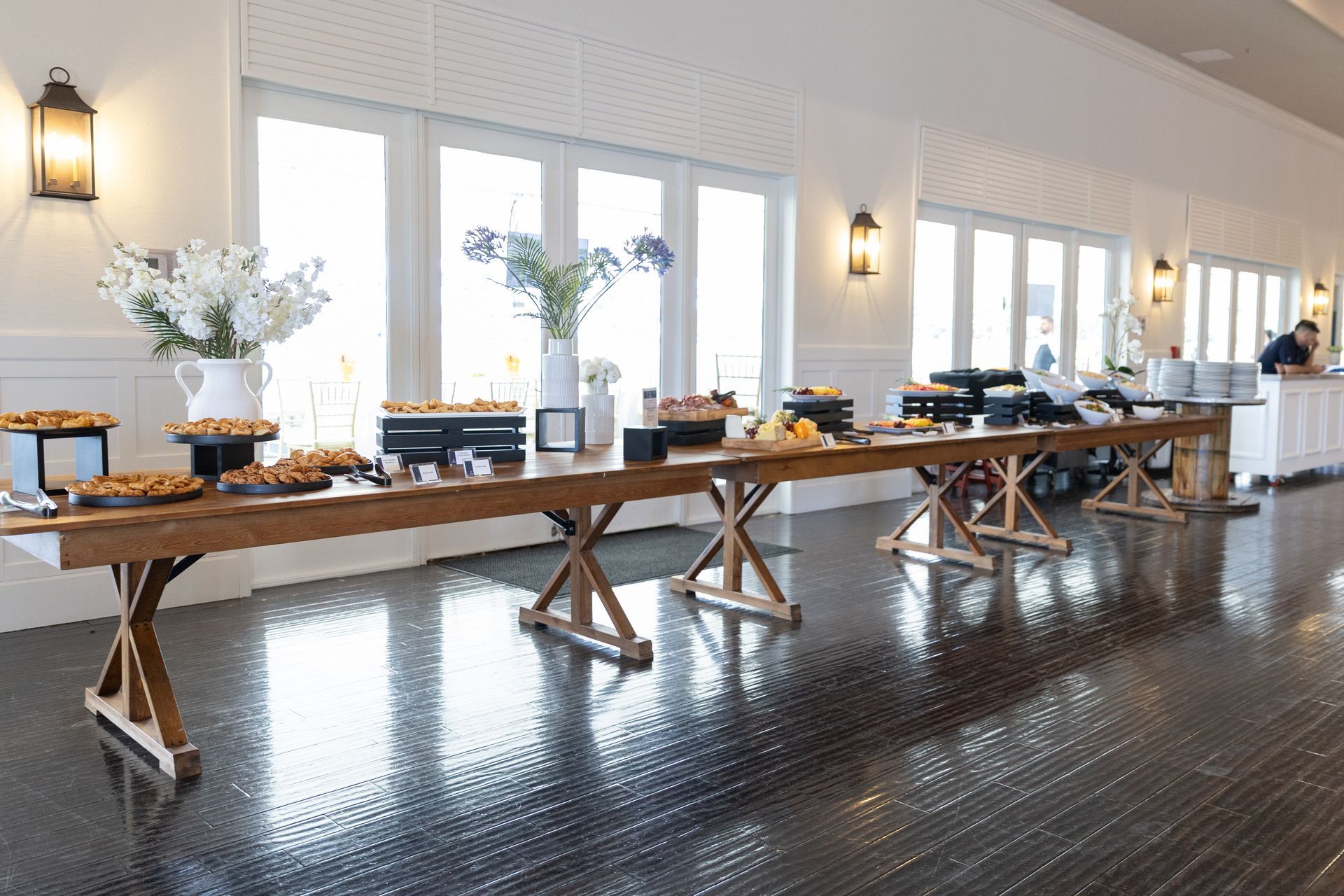 Long wooden buffet table with food and floral arrangements in a bright room with large windows.