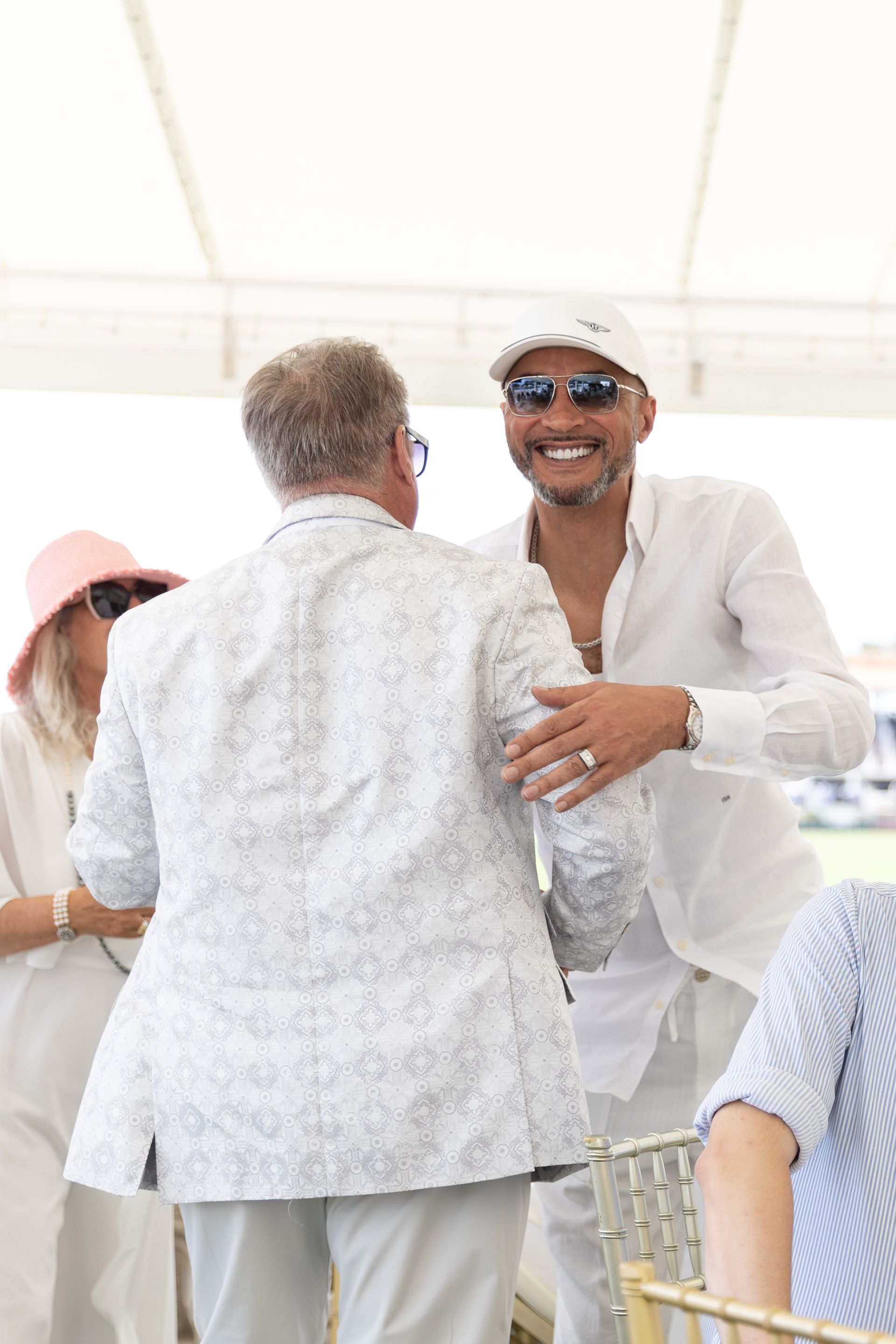 Two men in white outfits embrace outdoors, smiling. One wears sunglasses, a white cap.