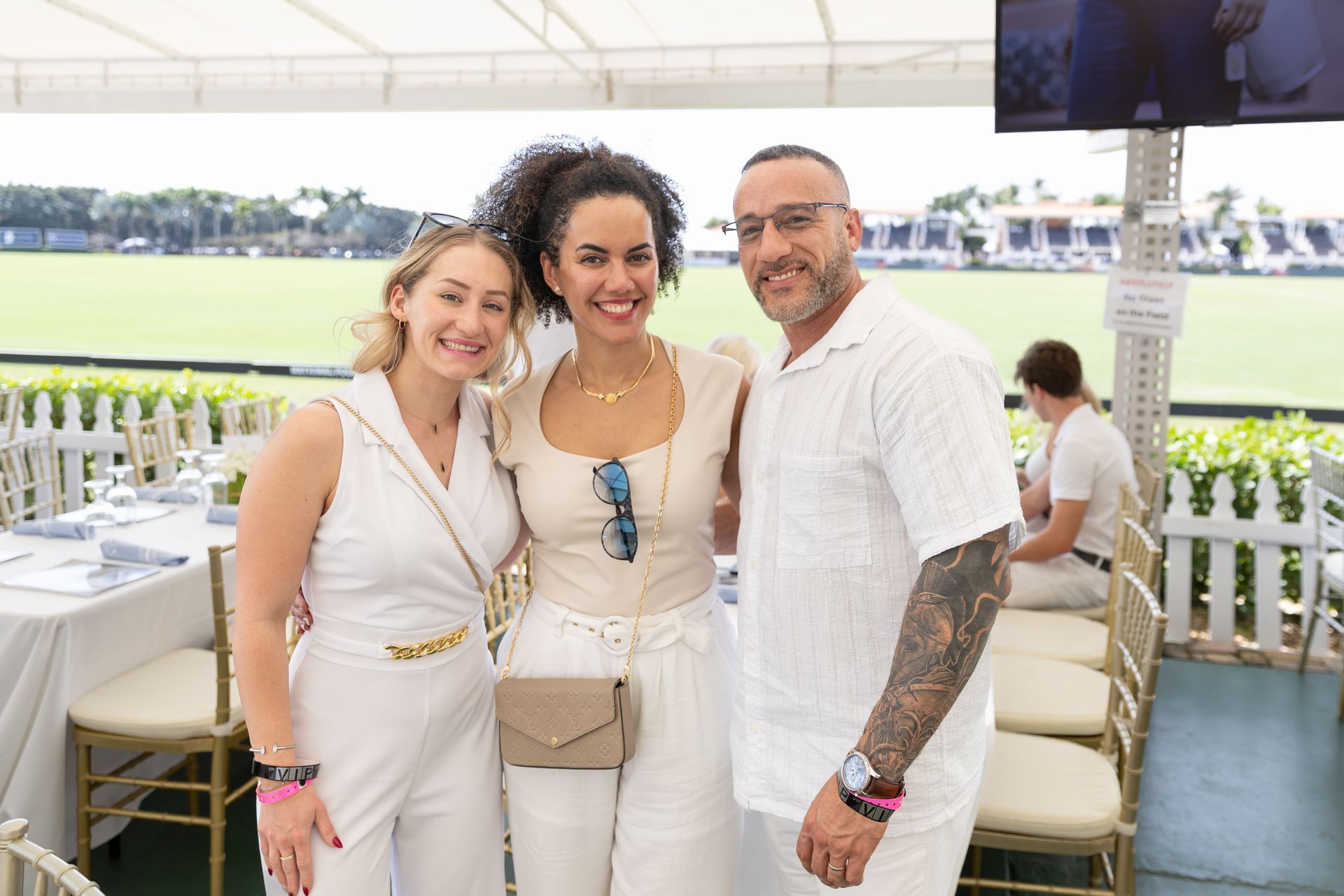 Three people in white outfits pose at an outdoor event with a field and tables in the background.