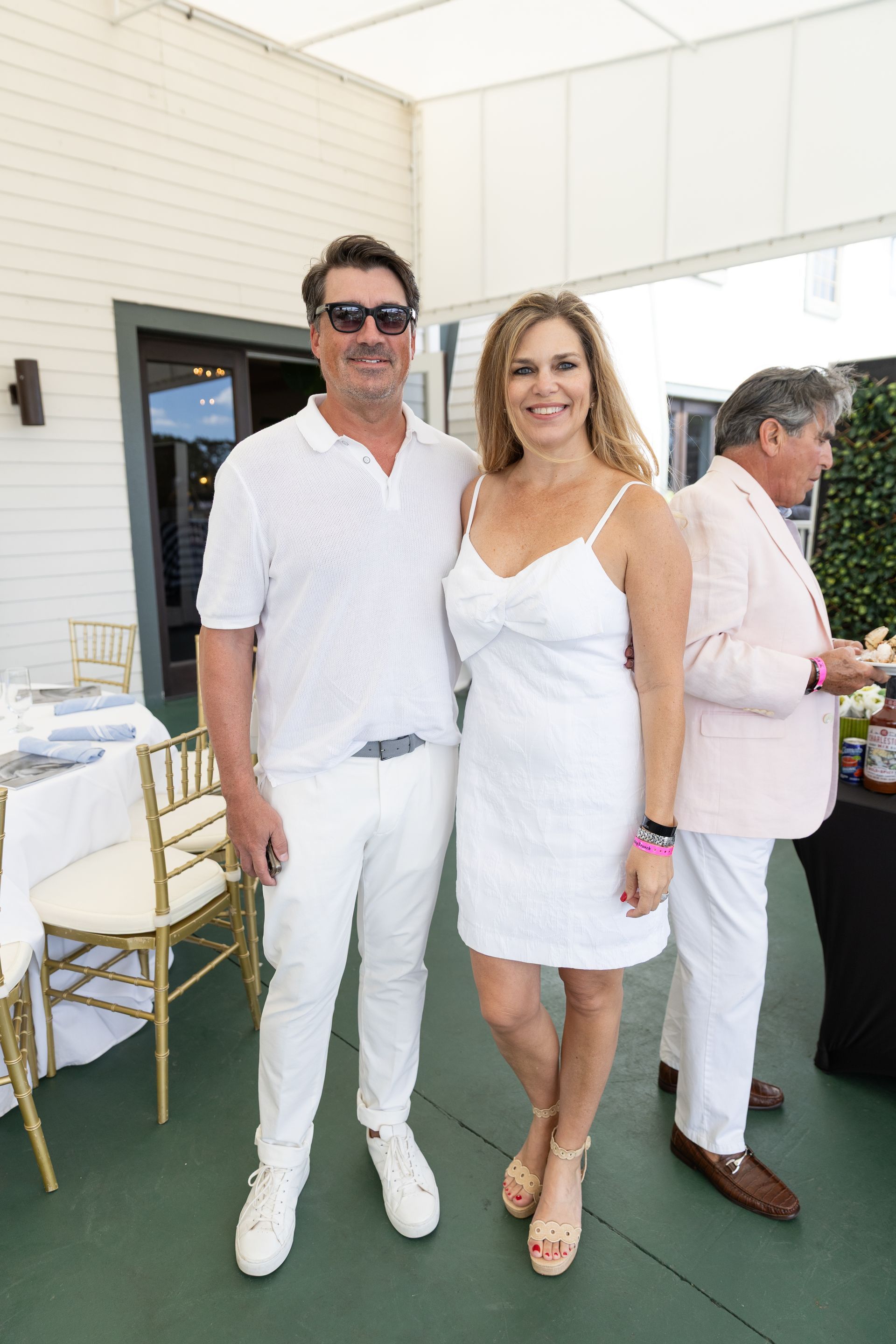 Couple in white outfits posing outdoors; man in polo and pants, woman in dress and heels.
