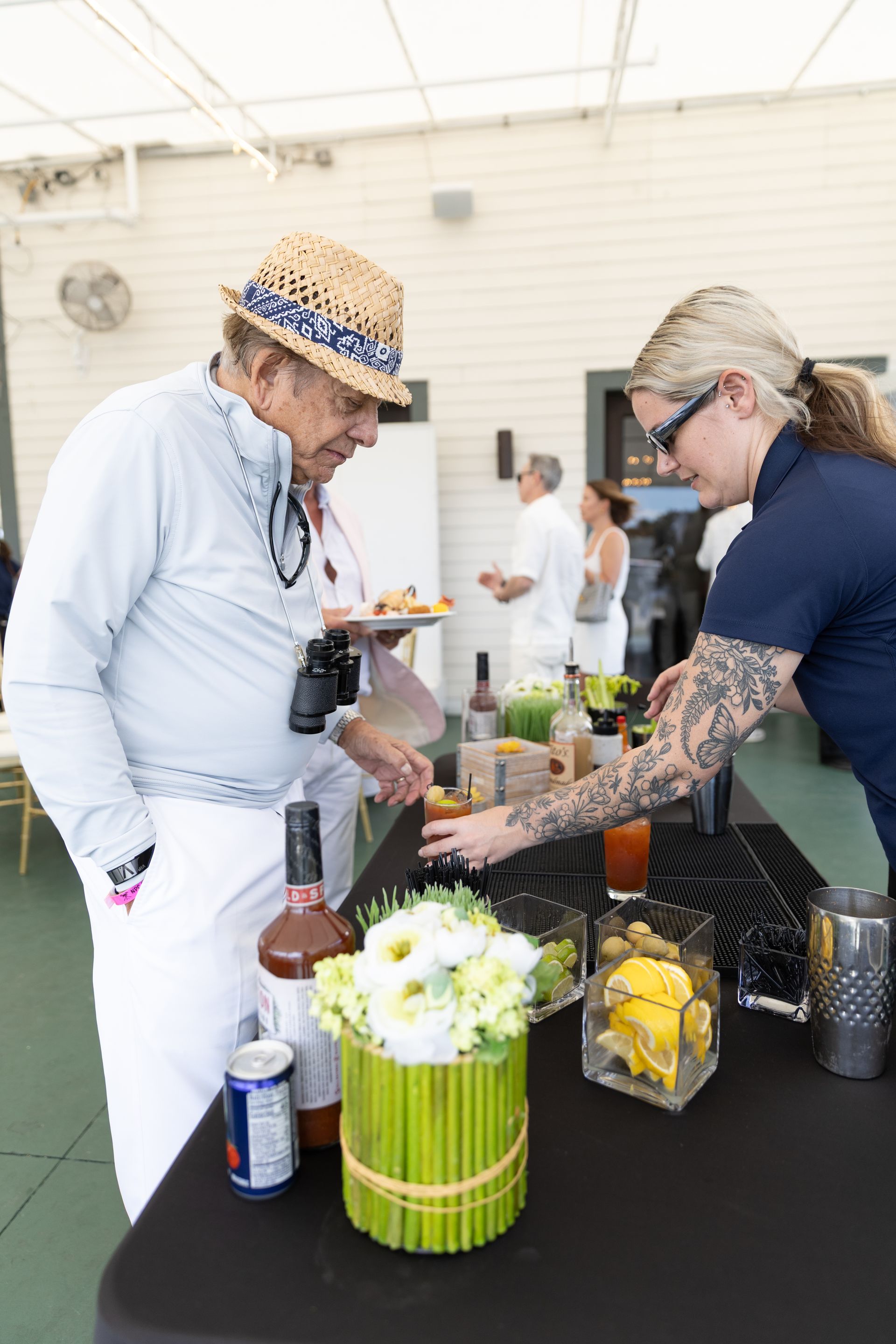 Man at a bar being served food by a woman with arm tattoos; setting is outdoors, table has drinks and flowers.