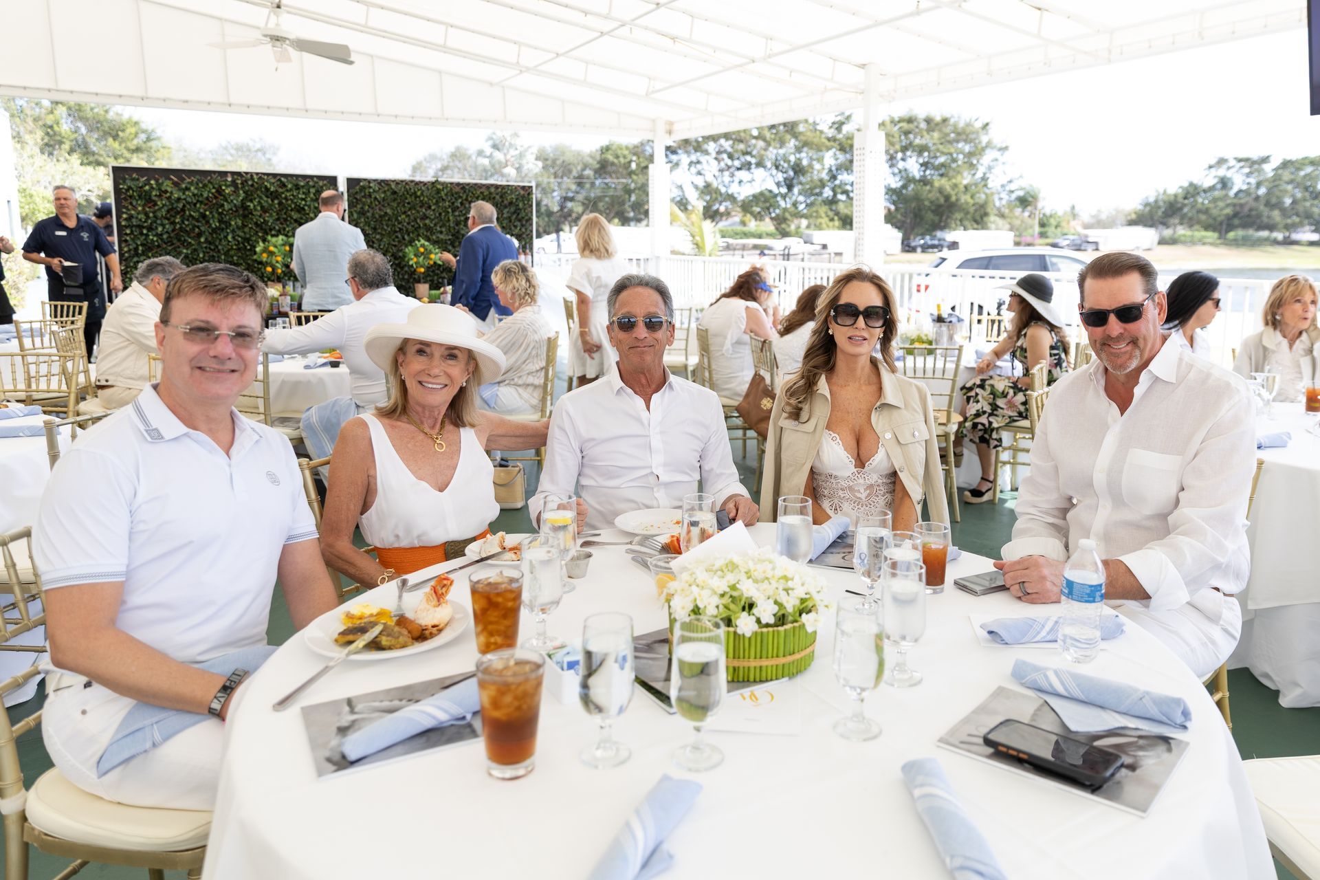 People at a table under a white tent; they're dressed in white and enjoying a meal with drinks.