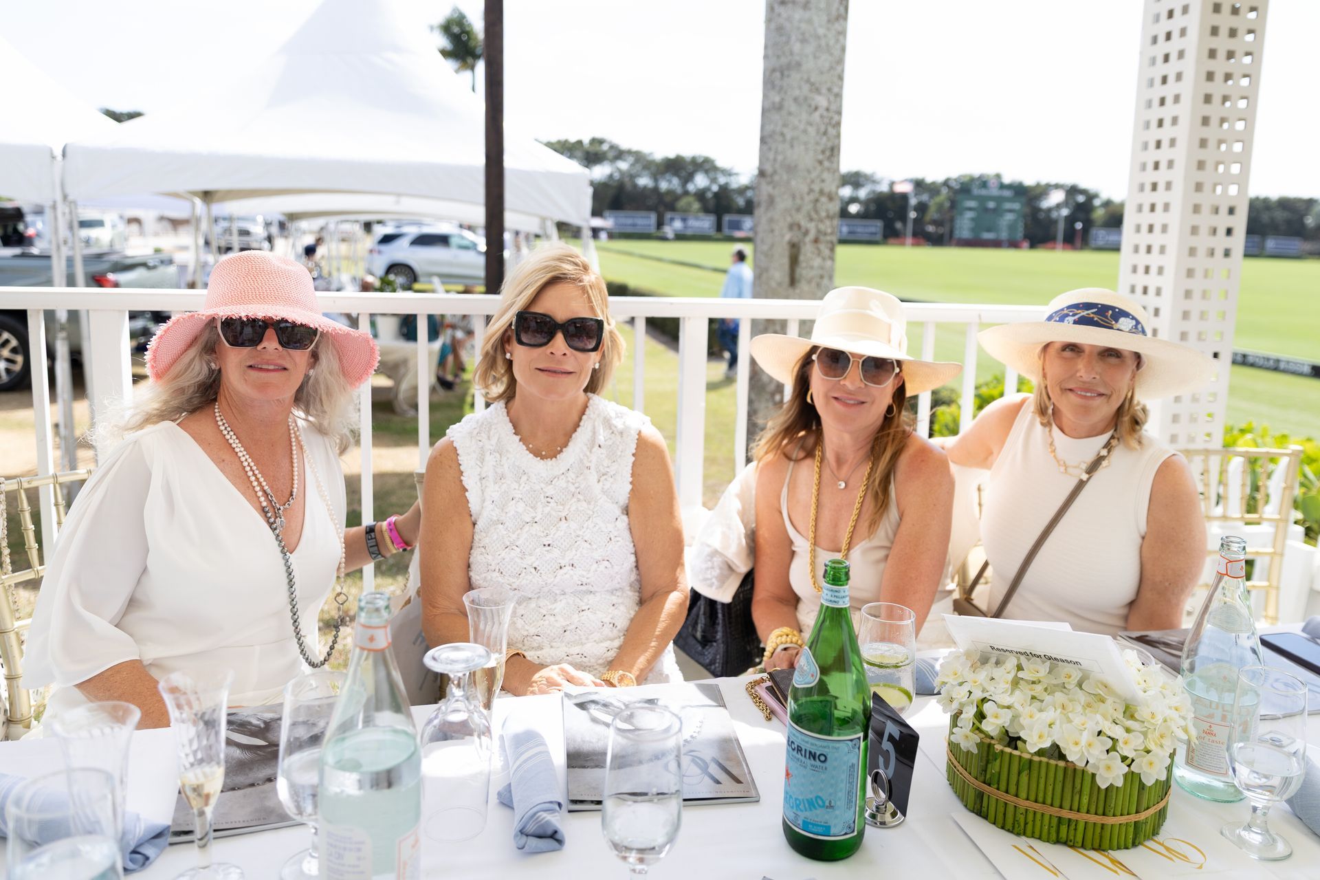 Four women in white outfits and hats seated at a table outdoors, with drinks.