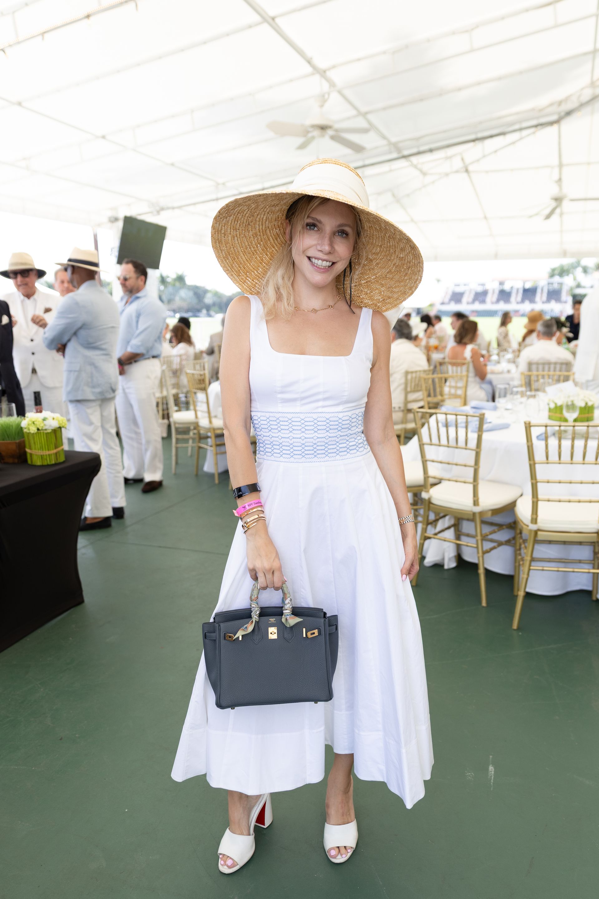 Woman in white dress and large hat, holding a handbag, at an outdoor event.