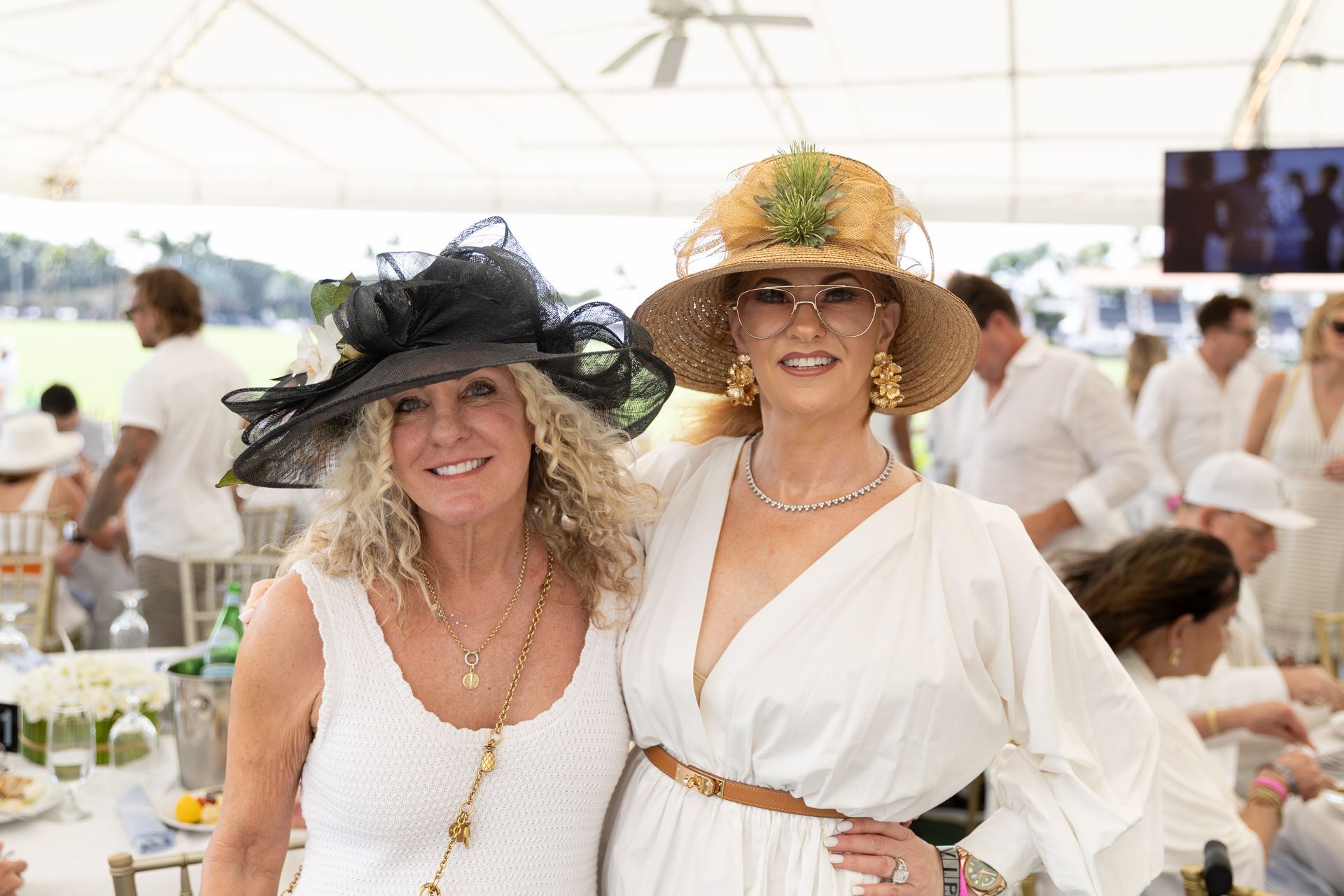 Two women in white outfits and hats smile at a polo event; people, tents and tables in the background.