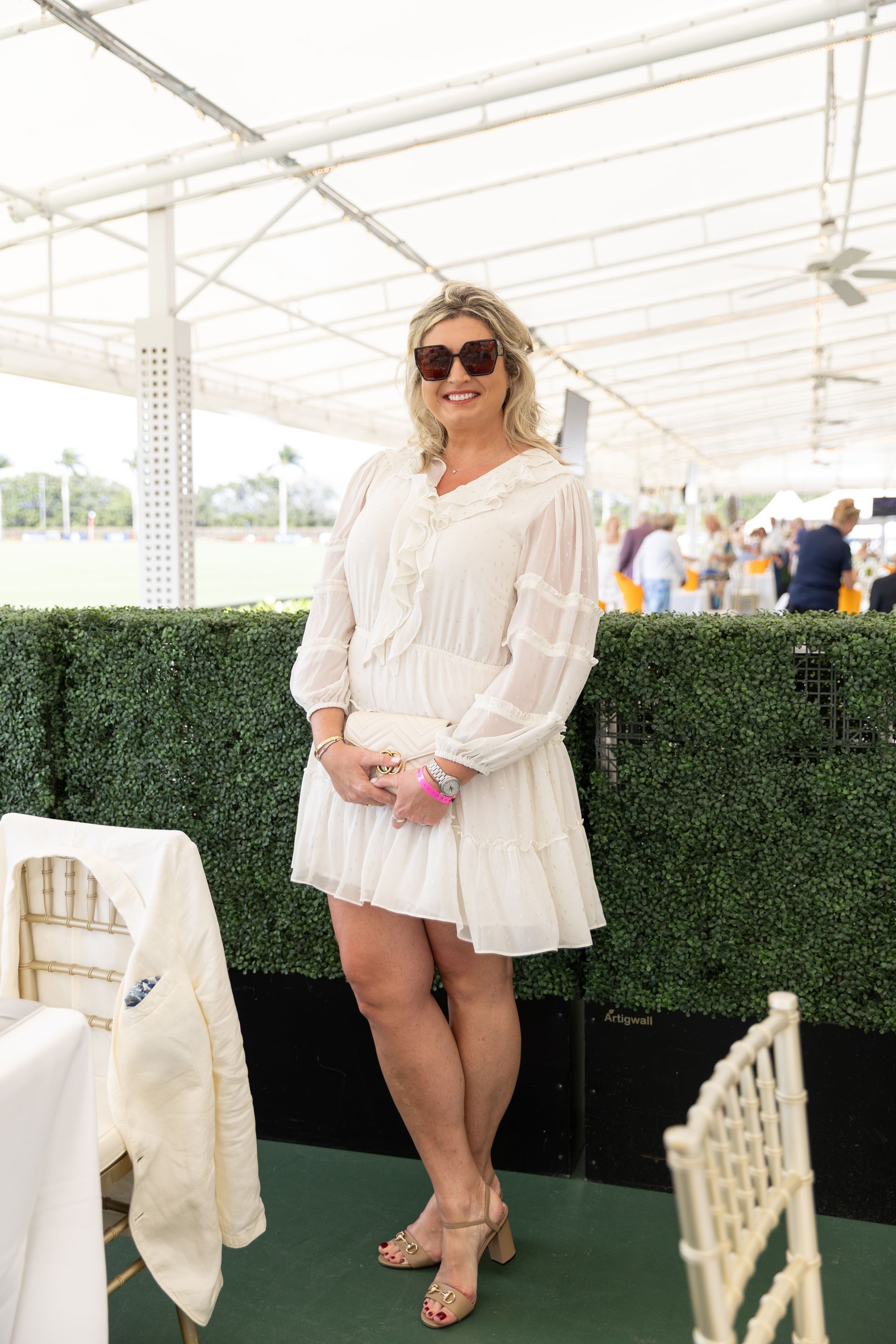 Woman in white dress and sunglasses at an outdoor event, holding a clutch.
