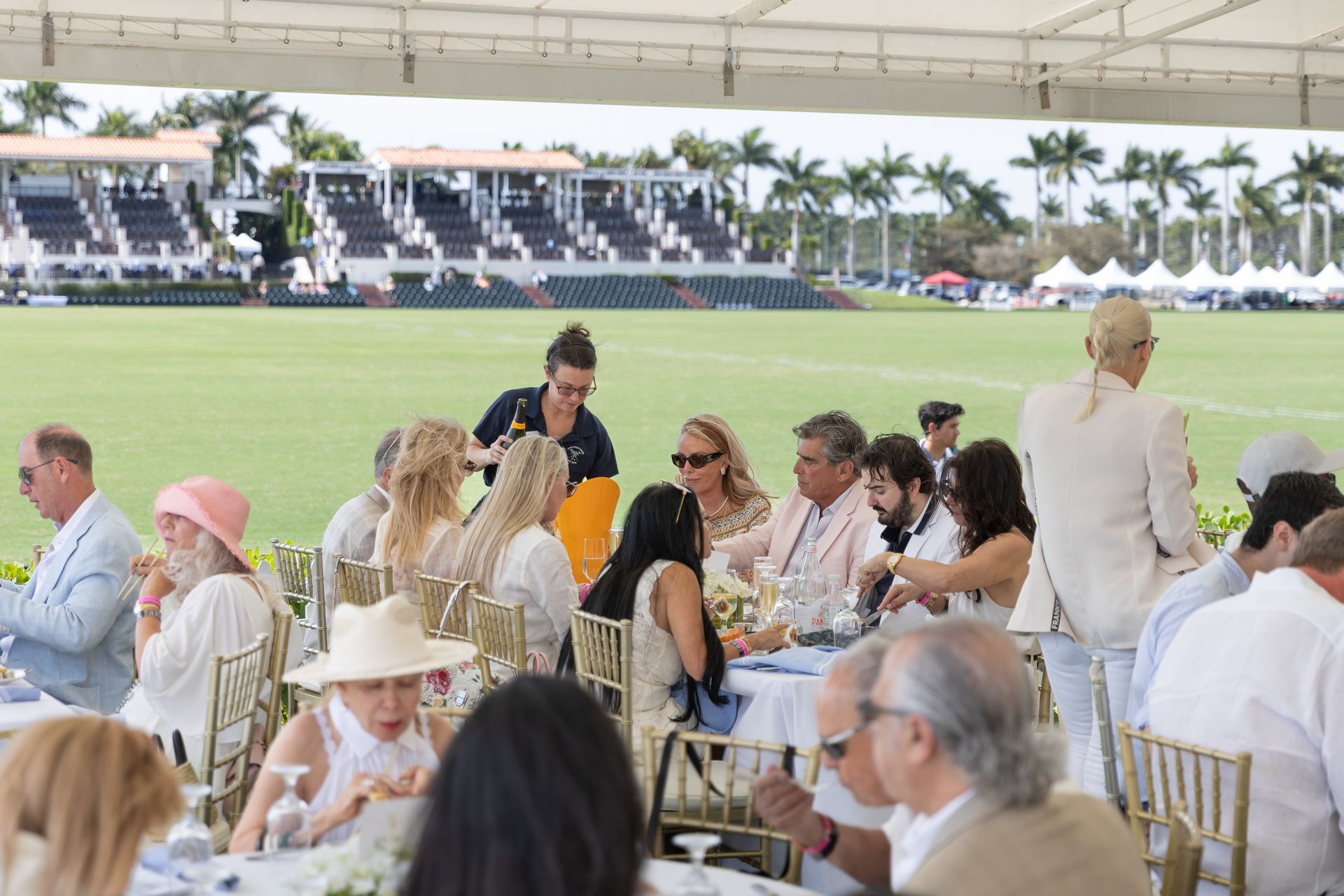 People dining at an outdoor event near a polo field; server pouring a drink.