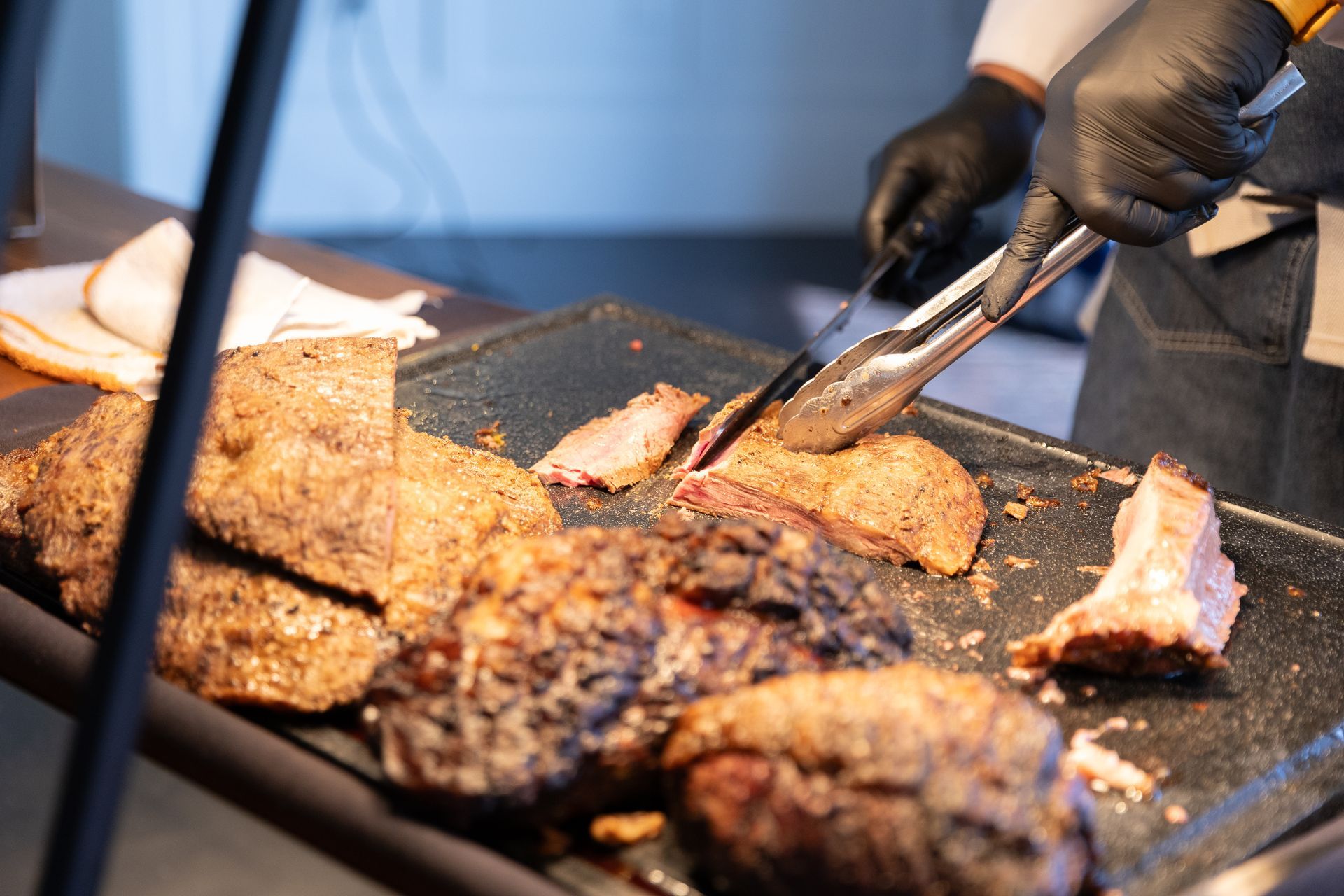 A person wearing black gloves using tongs to slice cooked meat on a tray.