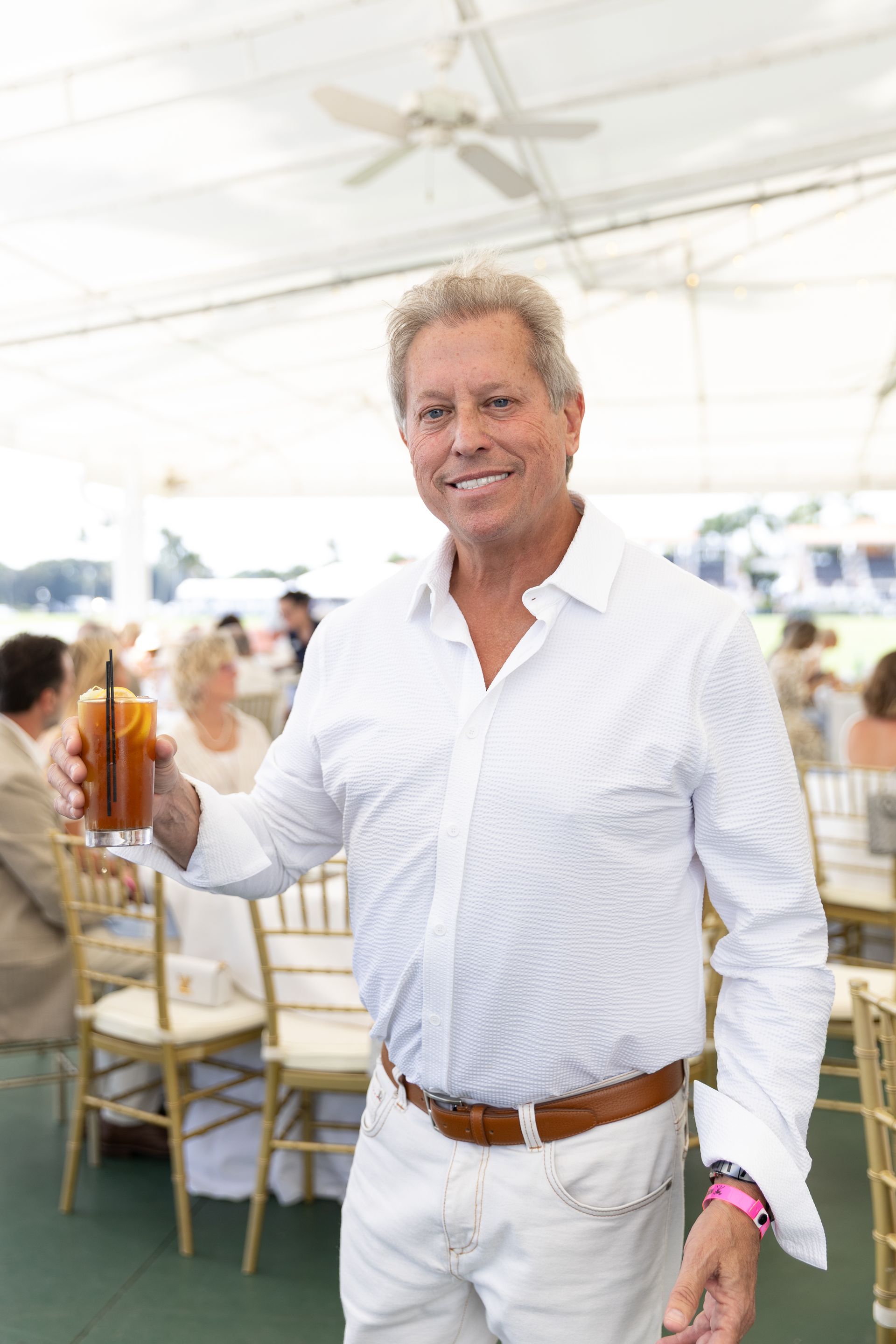 Man in white shirt and pants, holding a drink, smiling. Outside in a tent setting.