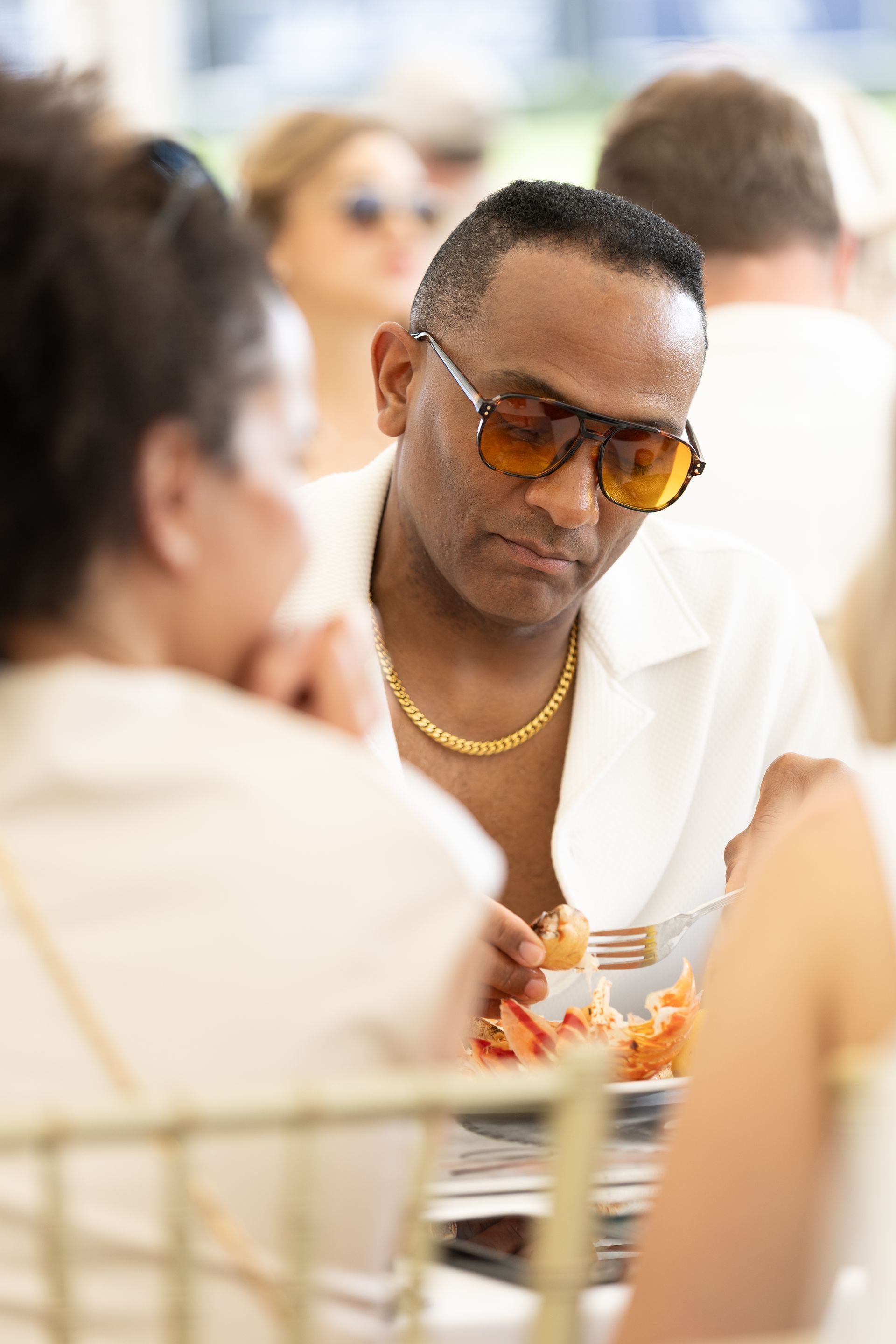 Man wearing sunglasses and gold chain, eating at a table, sunny outdoor setting.