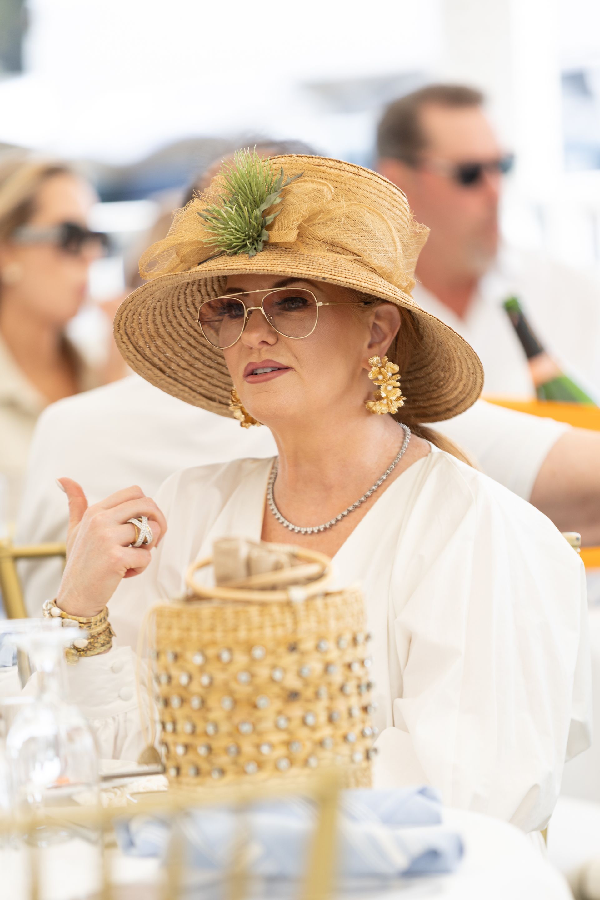 Woman in straw hat, sunglasses, and white outfit, seated at a table.