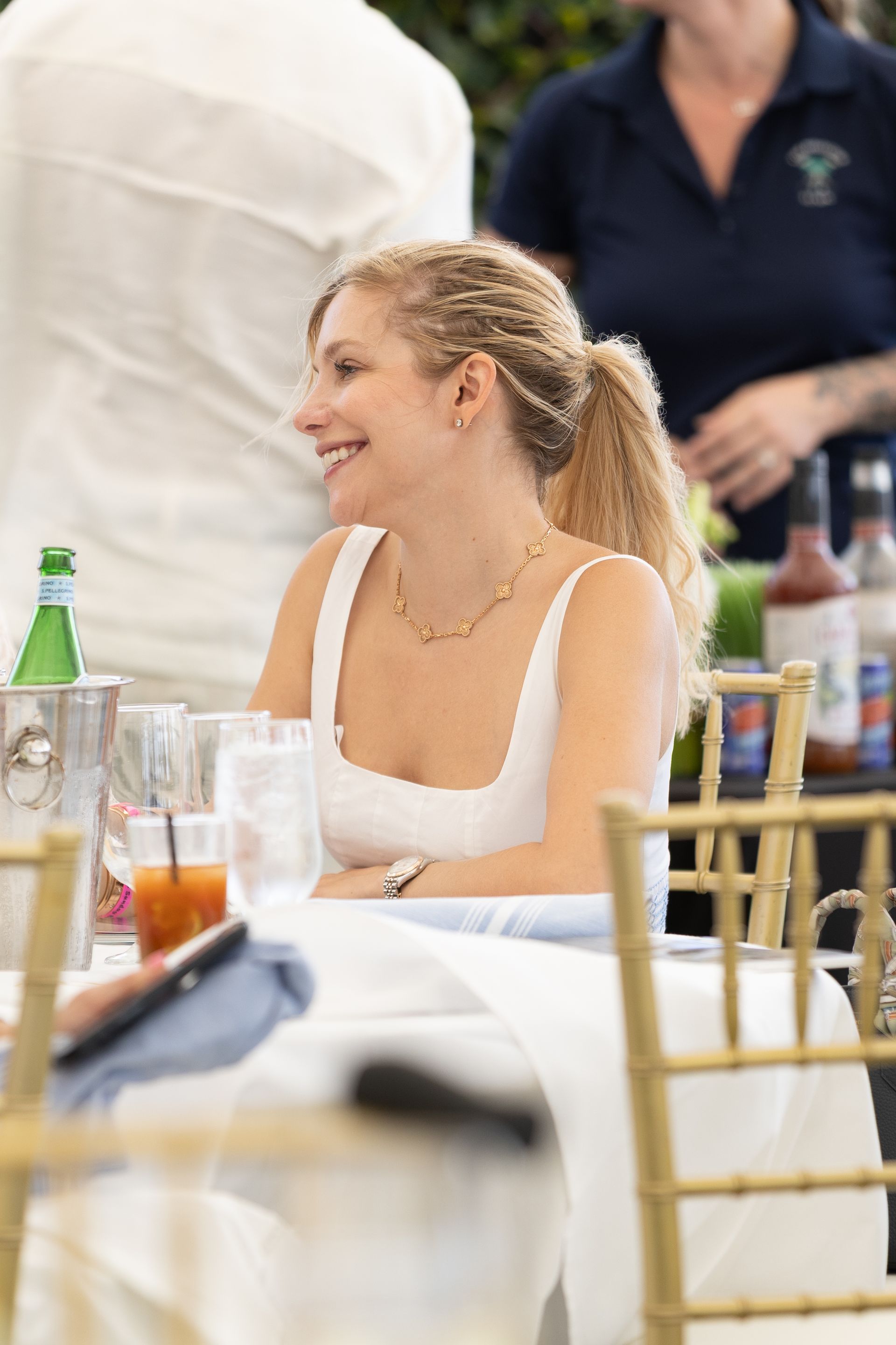 Woman with blonde hair smiles at a table outdoors. She wears a white top and a necklace.