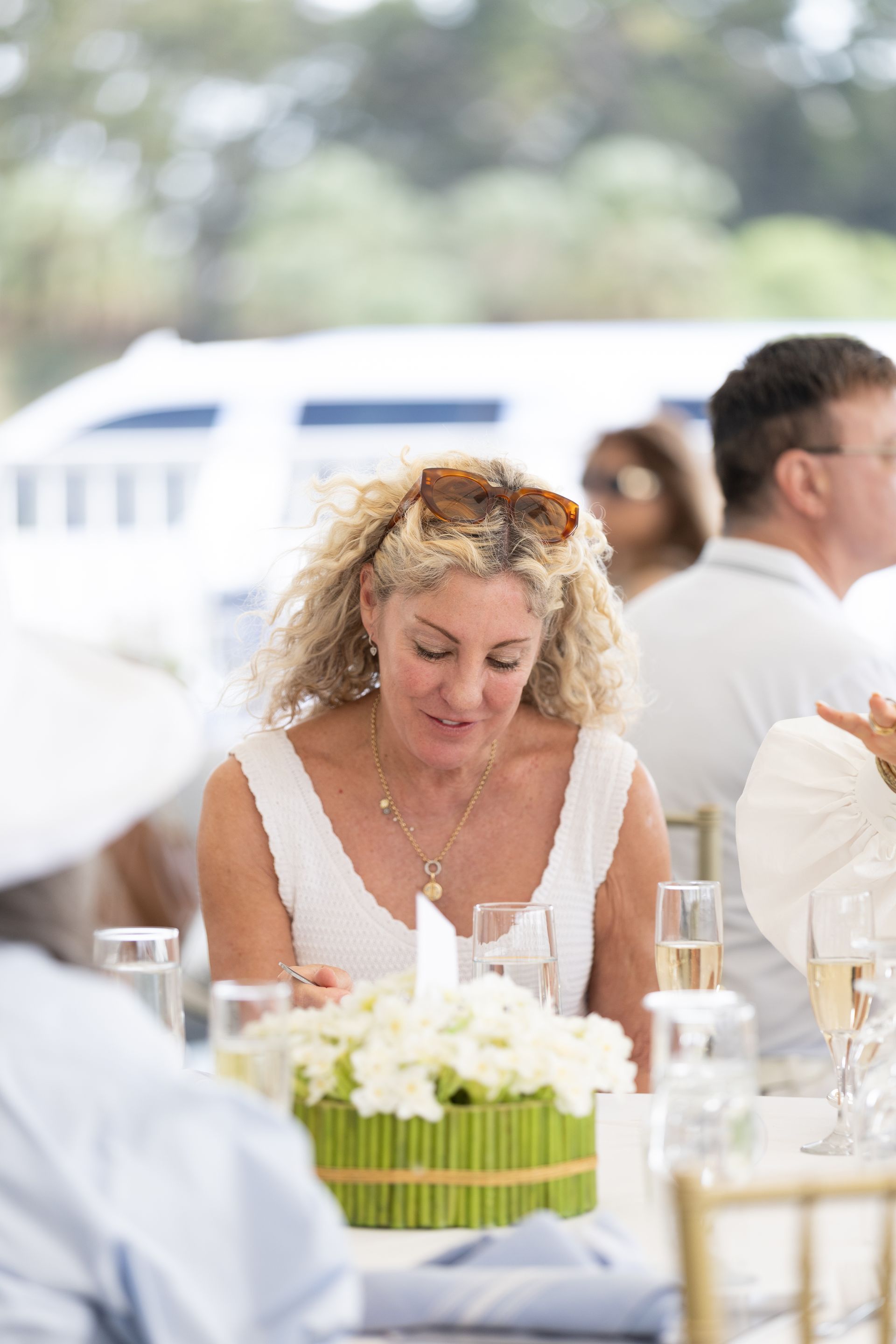 Woman at a table, wearing a white dress, blonde curly hair, sunglasses on head