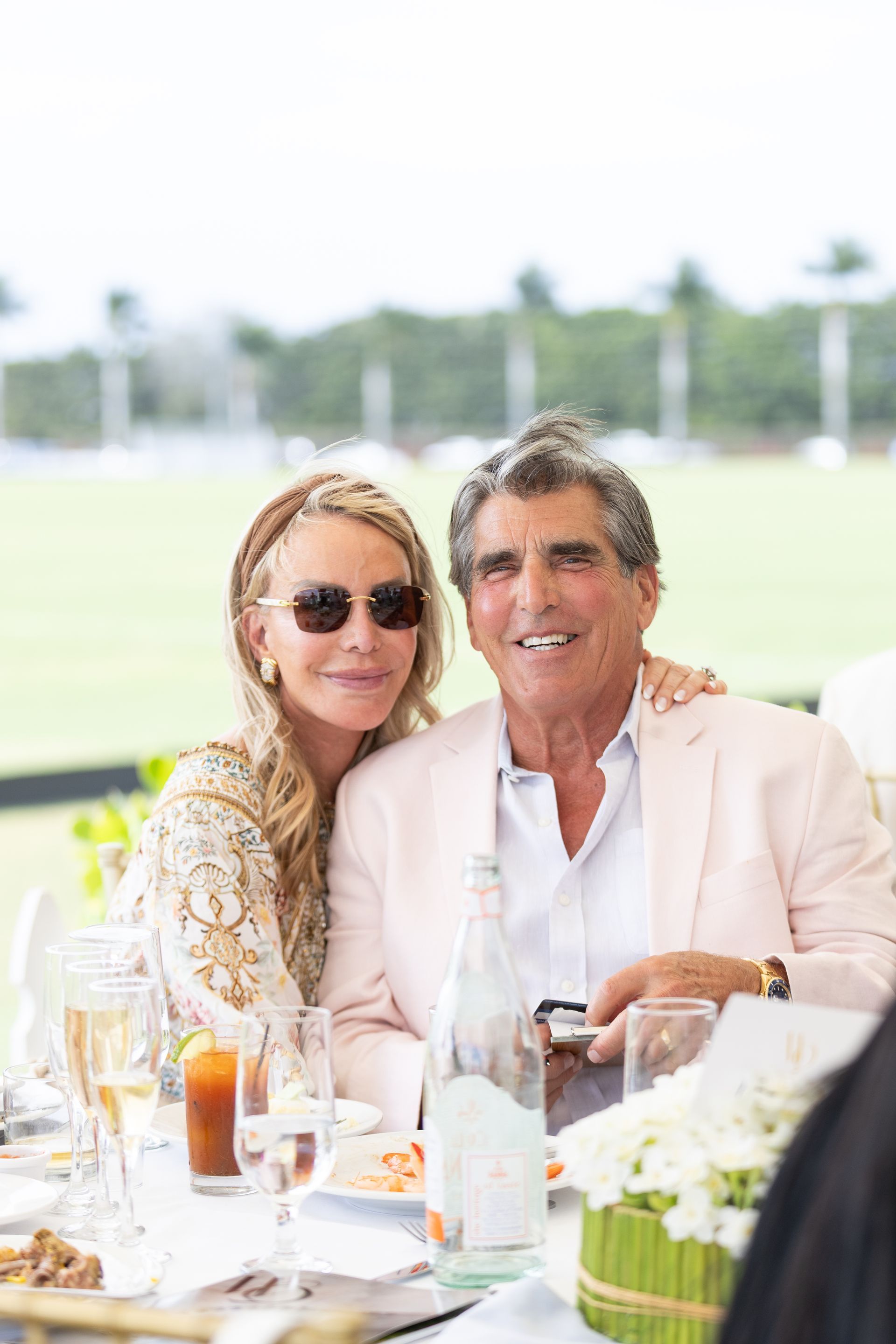 Couple at outdoor event, smiling. Woman in sunglasses, man in pink jacket, seated at a table.