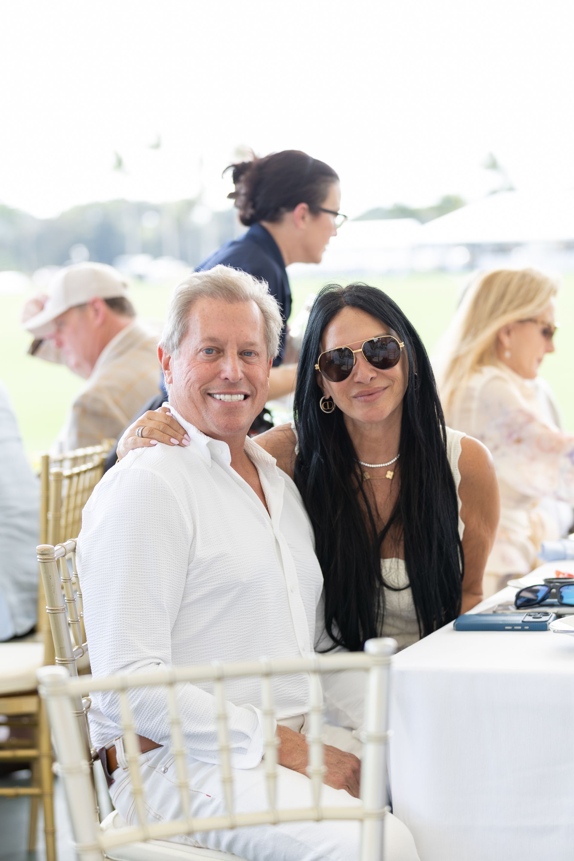 Man in white shirt with arm around woman with long dark hair, both smiling, sitting outdoors.