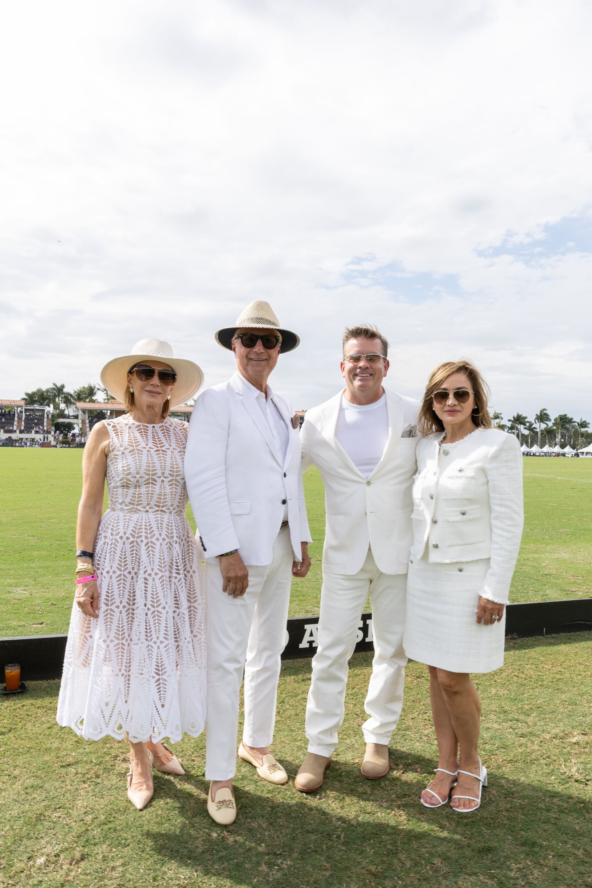 Four people in white attire pose outdoors on a grassy field; sunny day.