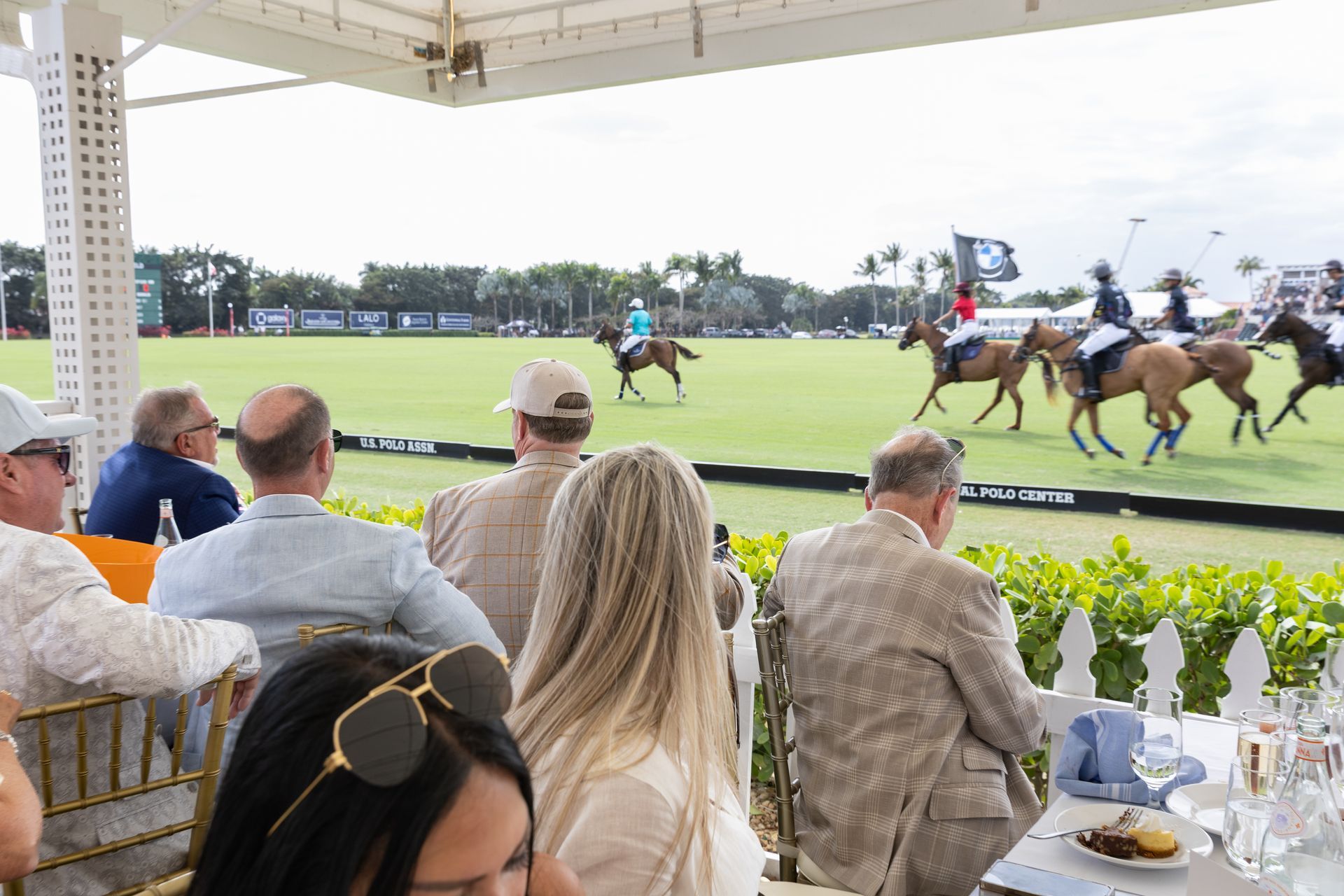 Spectators watch a polo match from a covered outdoor seating area. Horses and players on a green field.