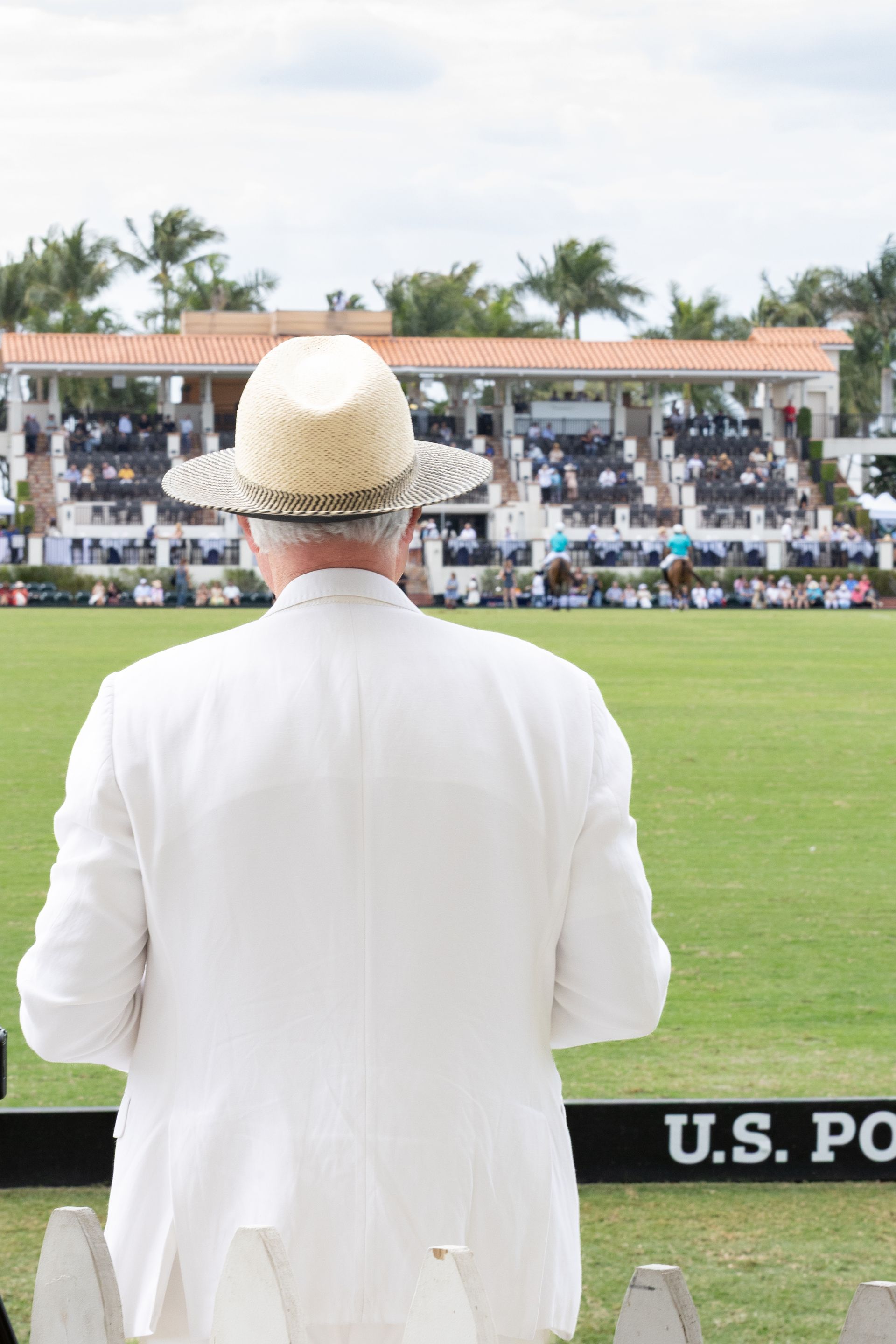 Man in white suit and straw hat watches a polo match, stands in front of the stands and field.