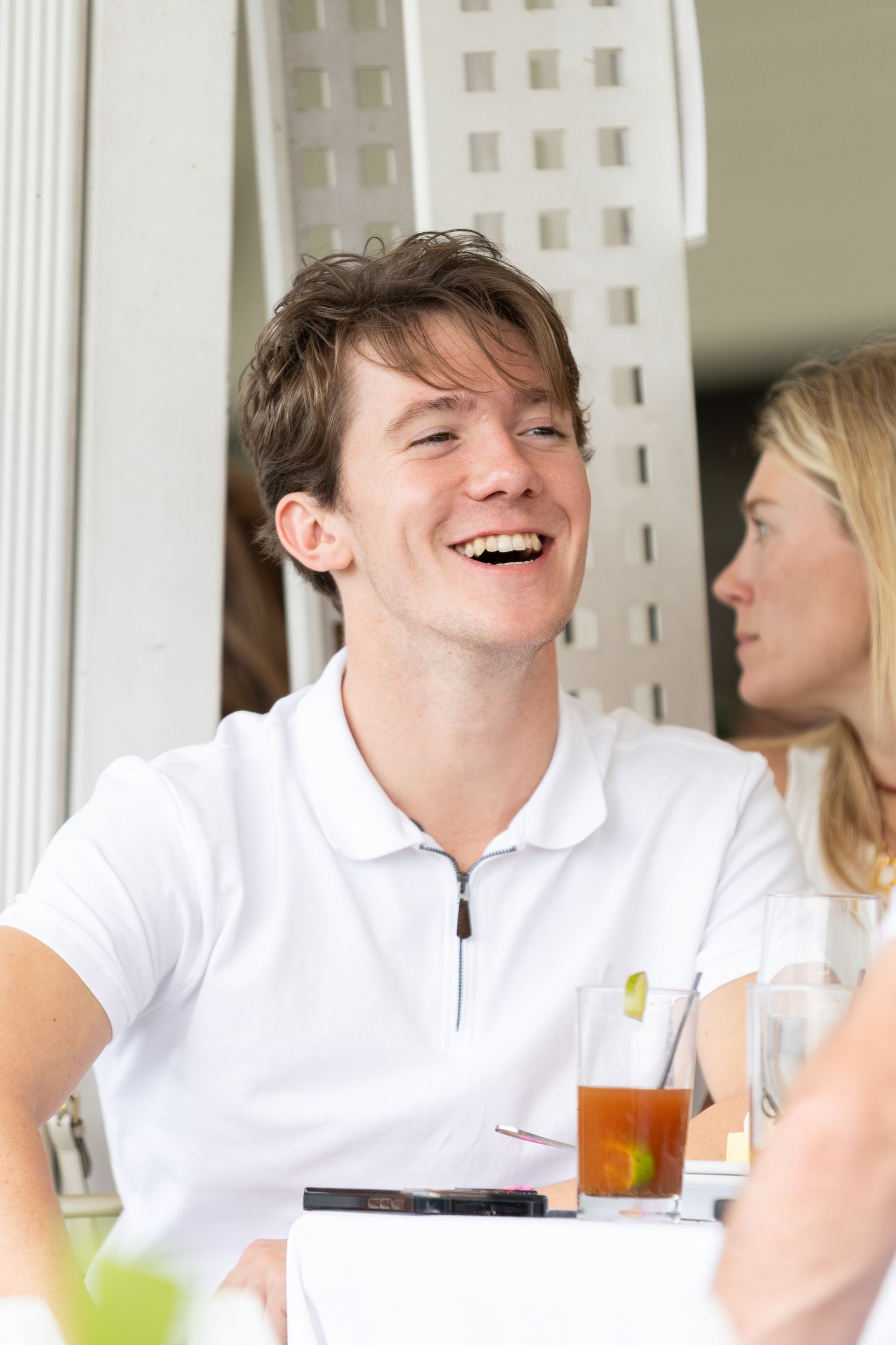 Man in white polo shirt laughs, sitting at a table with a drink and another person visible.