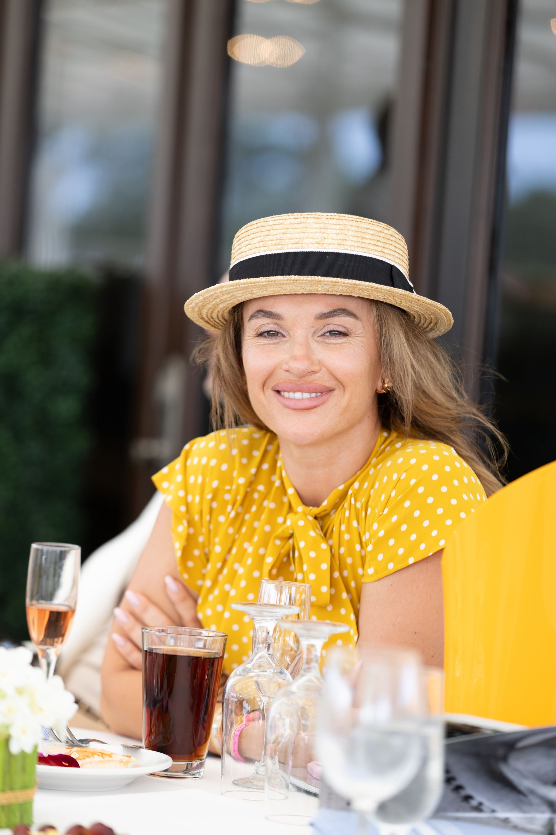 Woman wearing a straw hat and yellow polka dot blouse smiles at outdoor table with drinks.