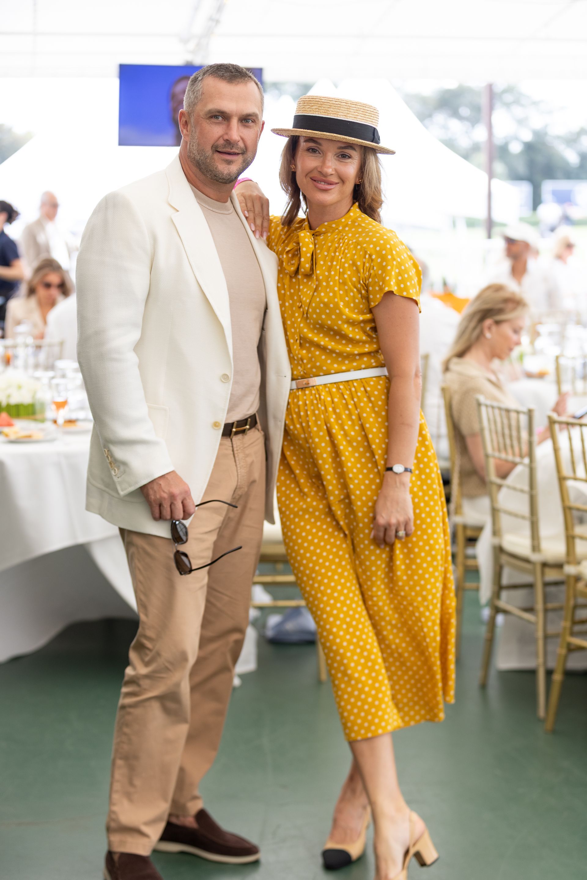 A couple poses at an outdoor event. Woman in yellow polka-dot dress, man in cream blazer.