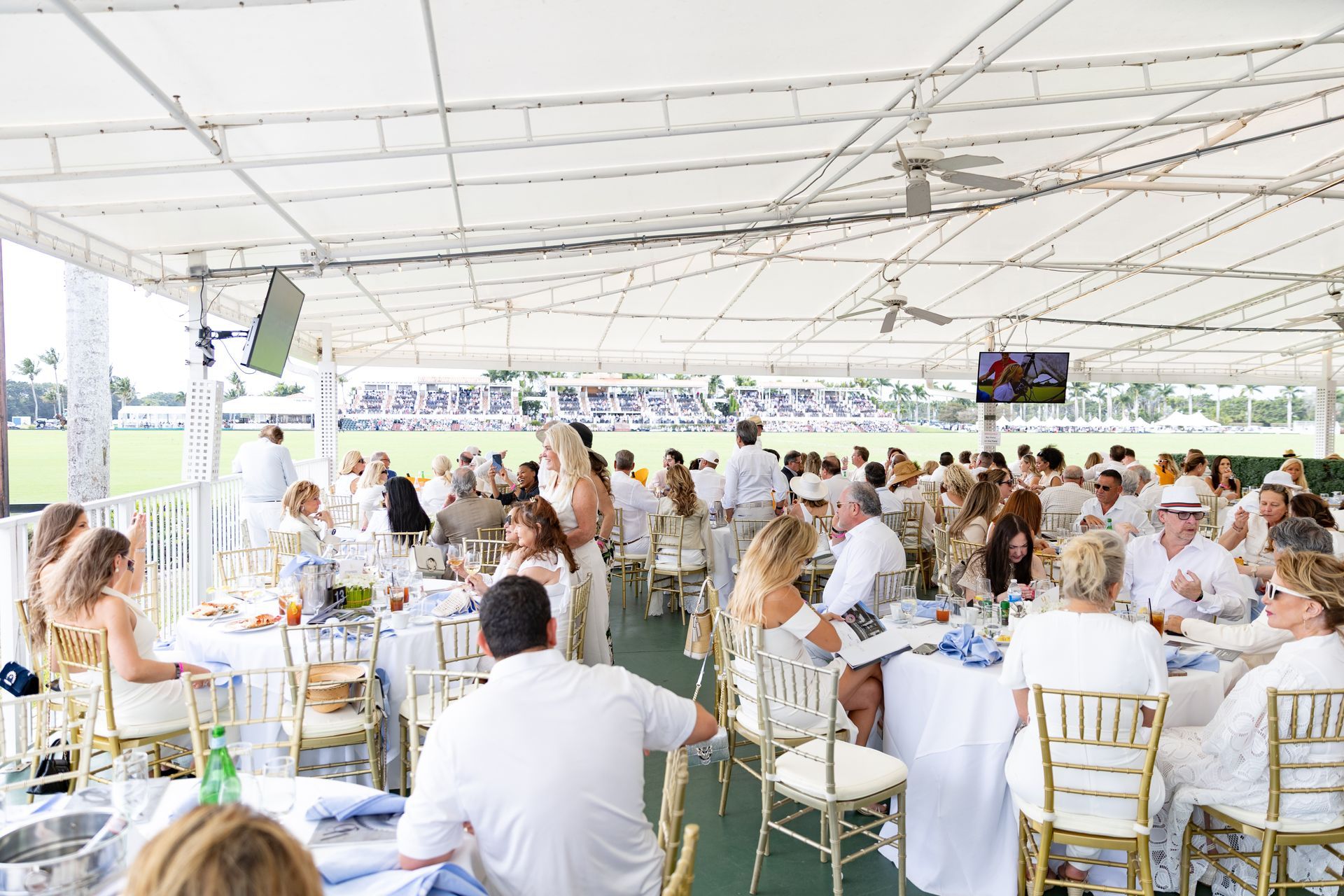 People dining at tables under a white canopy tent, overlooking a polo field.
