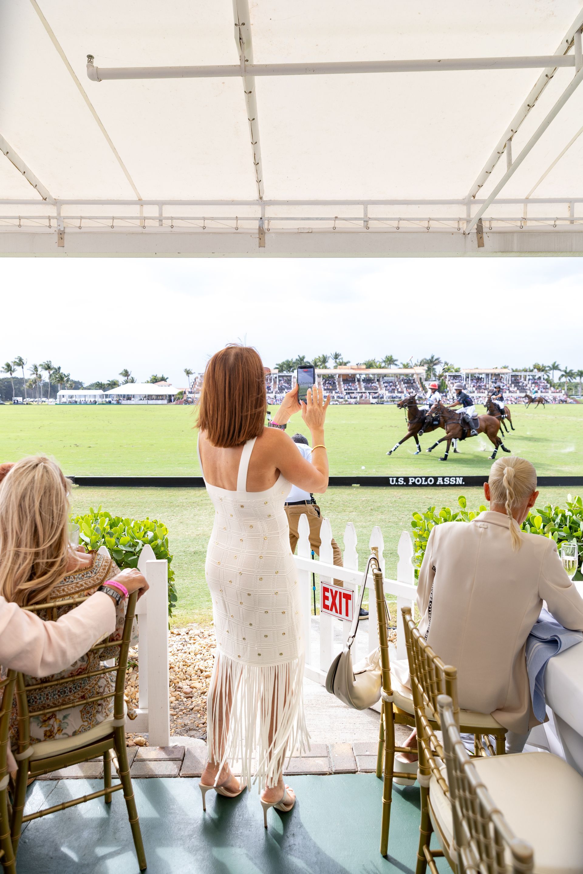 Woman in white dress photographs polo match. Spectators watch from under a canopy.