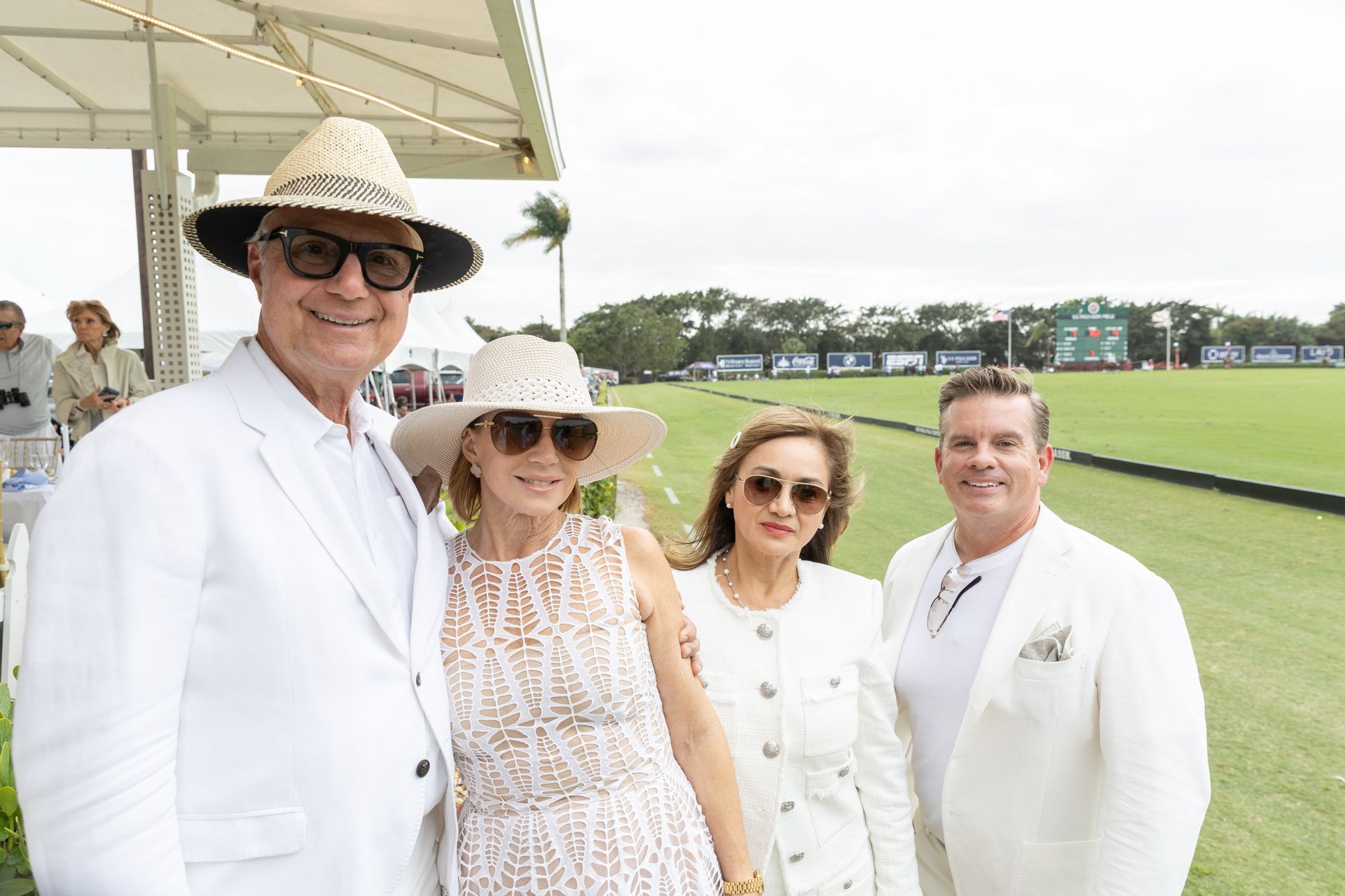 Four people in white attire pose outdoors at a polo match.