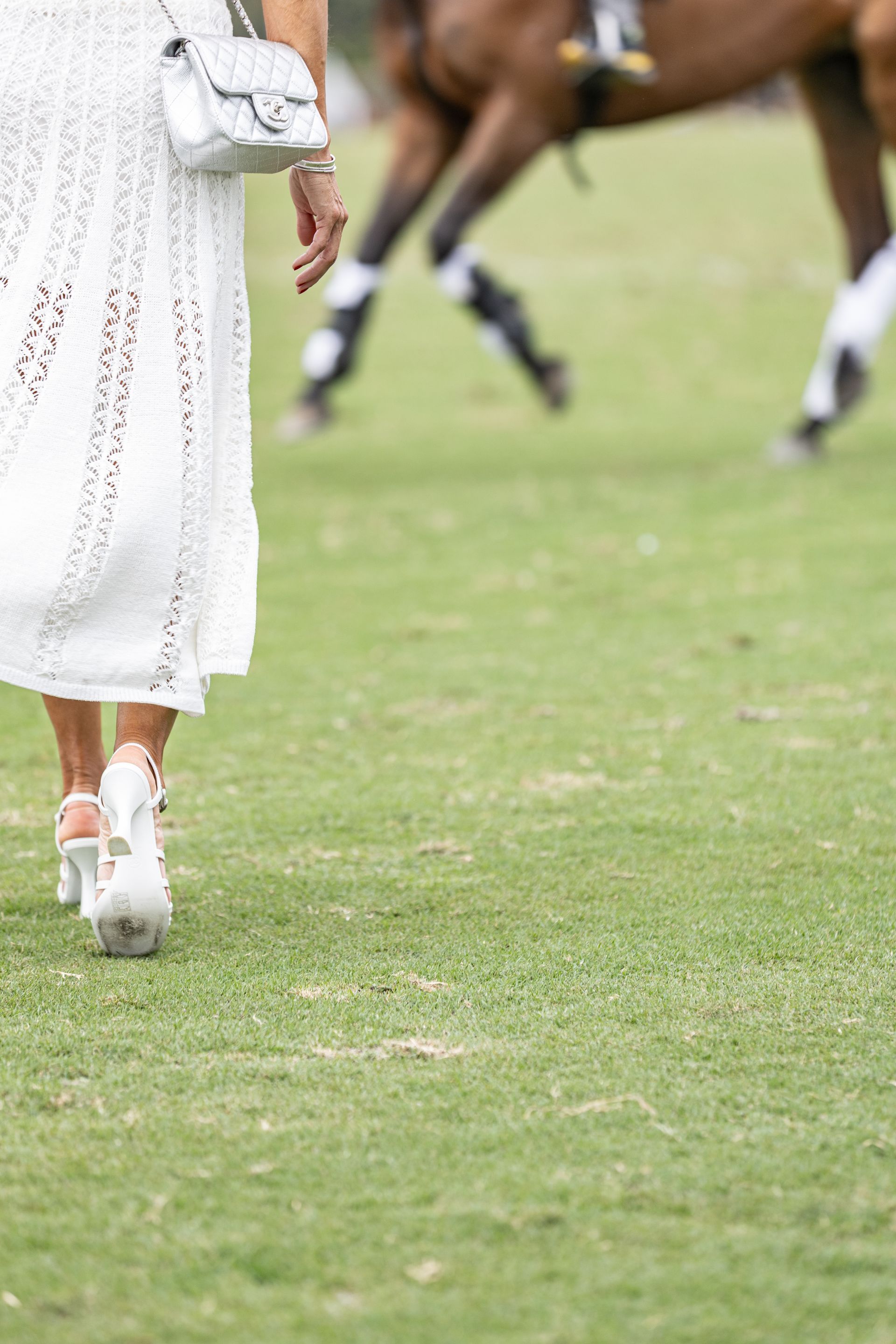 Woman in white dress and heels walks on grass, horse in the background.
