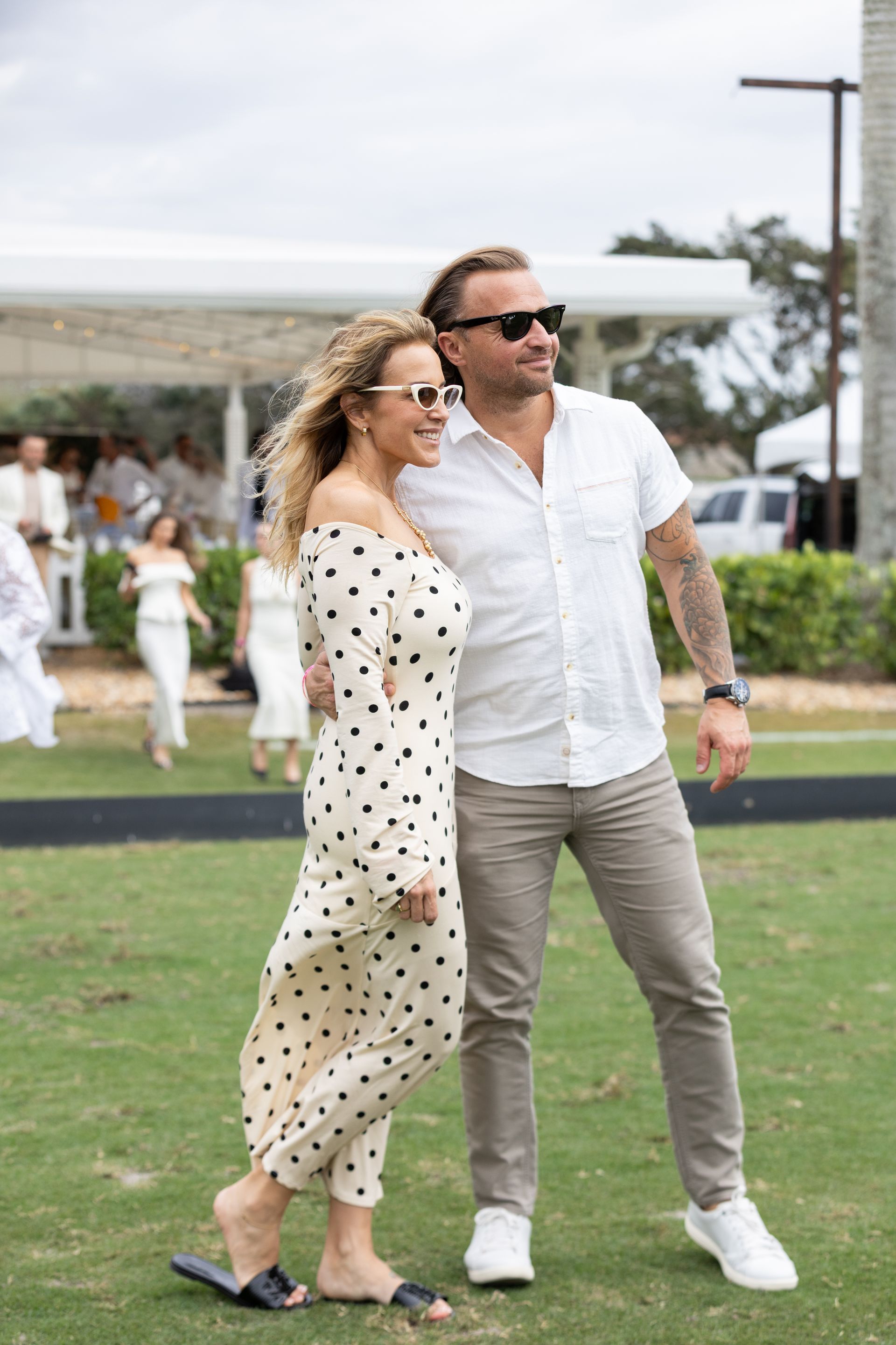 Couple poses outdoors: woman in polka-dot dress, man in white shirt and khaki pants, both with sunglasses.