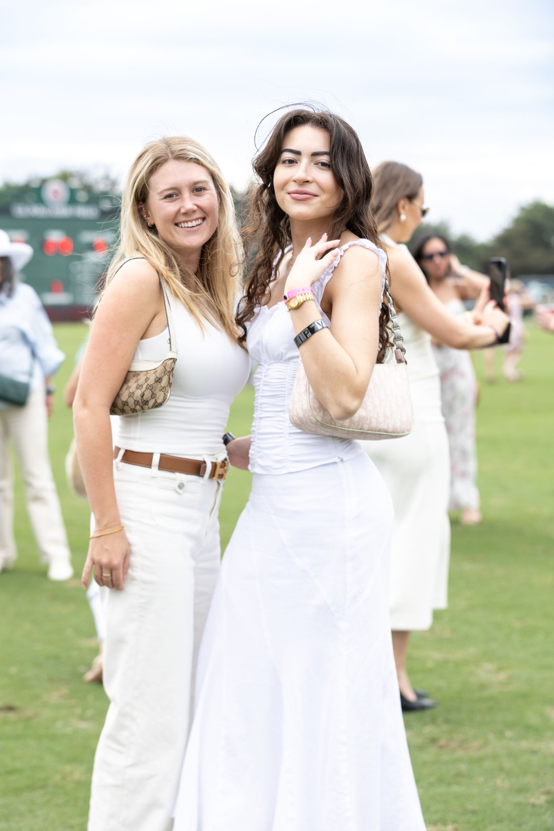 Two women smiling on a grassy field. One in white dress, holding a purse. The other wears white top and pants.