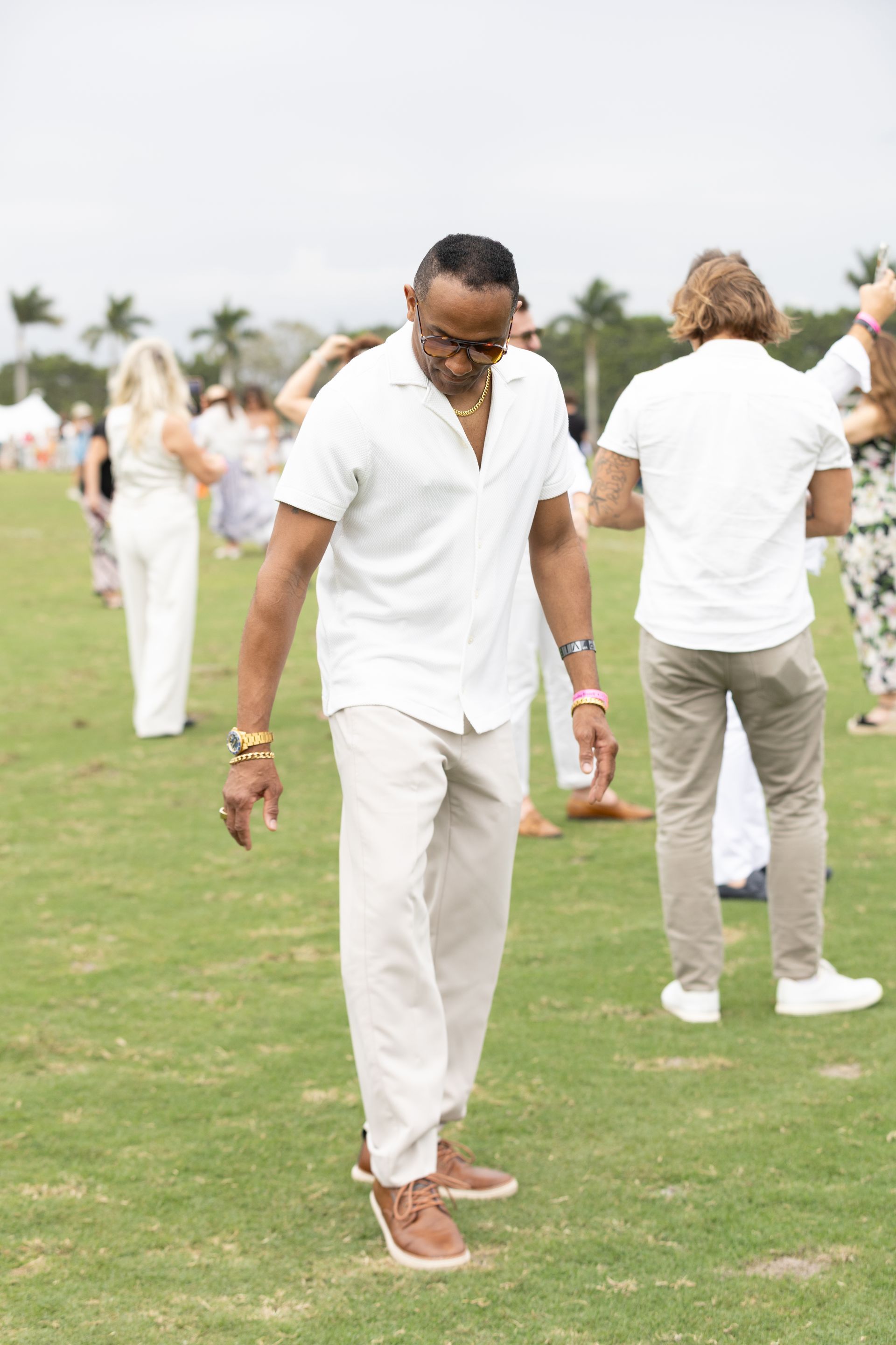 Man in white shirt and pants outdoors, looking down. Others in white clothing on a grassy field.