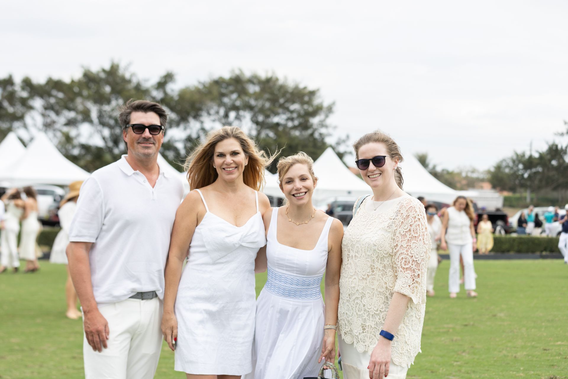 Group of people smiling, dressed in white, on a grassy field with tents in the background.