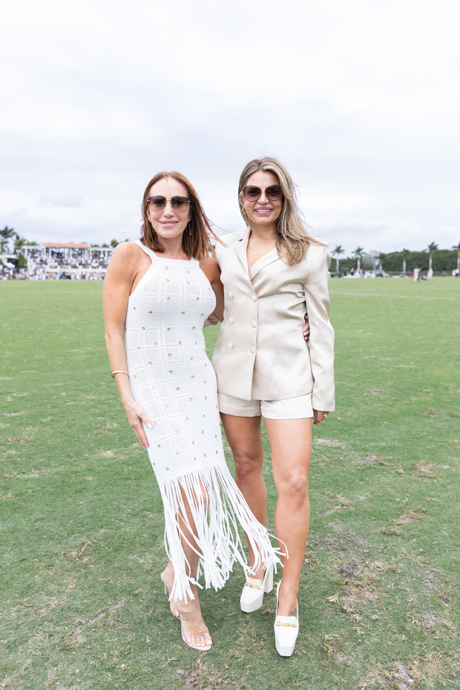 Two women pose outdoors: one in white fringe dress, the other in a tan shorts suit.