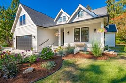 A light-gray, two-story house with a front porch, garage, and landscaped garden under a clear blue sky.