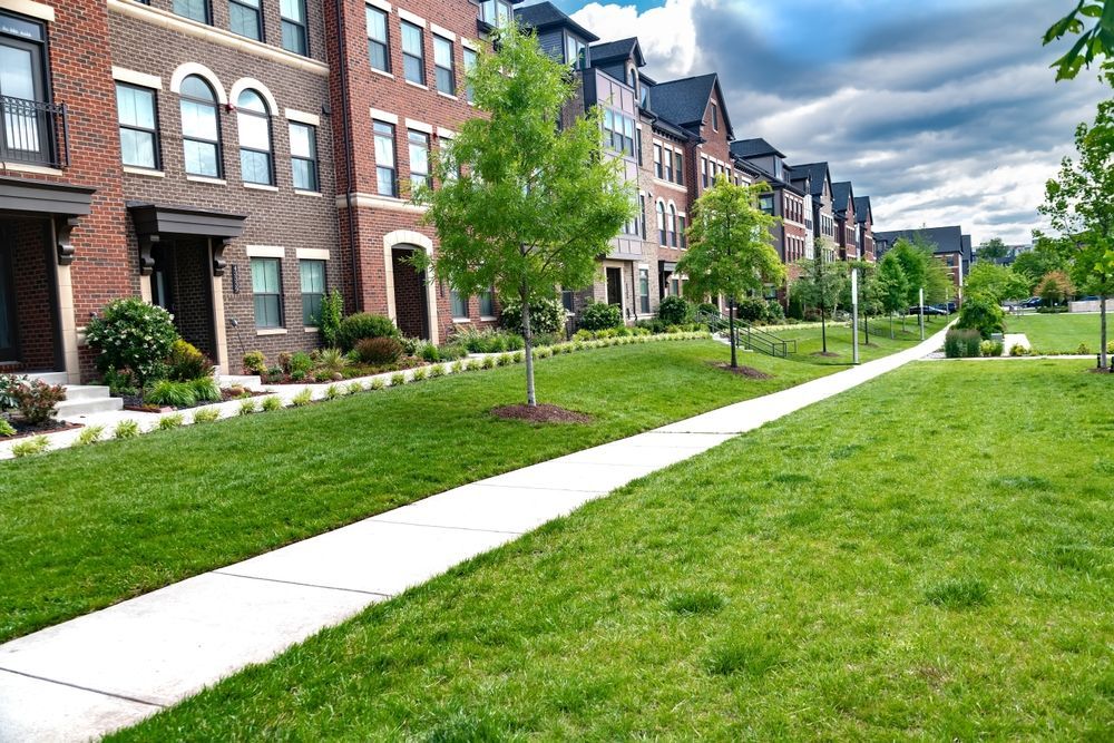 A row of multi-story brick townhouses with a sidewalk running alongside a grassy lawn under a cloudy sky.