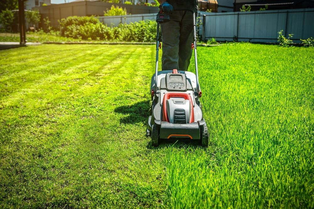 A person pushing a grey and orange lawn mower across a sunny green lawn, leaving a trail of neatly cut grass.
