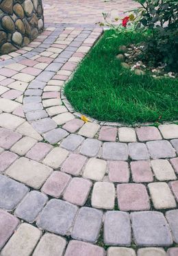 A curved stone paver walkway borders a small patch of green grass and a rose bush next to a stone-walled structure.